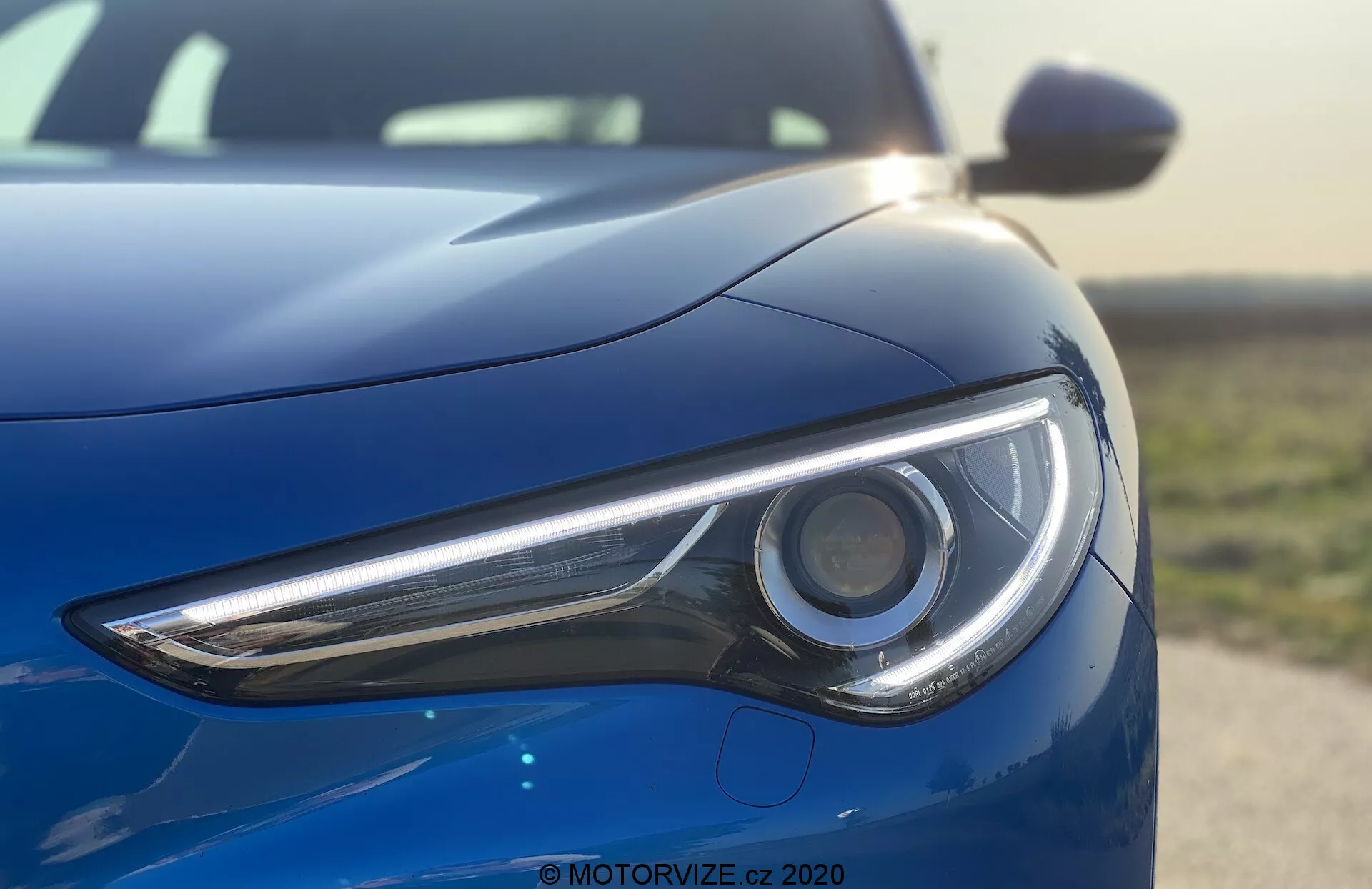 Vista aproximada da seção frontal esquerda do Alfa Romeo Stelvio 2017 destacando o farol, tirada de um ângulo baixo. A imagem enfatiza o design elegante e a tecnologia LED do conjunto de luzes do modelo de luxo esportivo com uma carroceria de alto brilho em um azul metálico profundo refletindo um céu limpo.