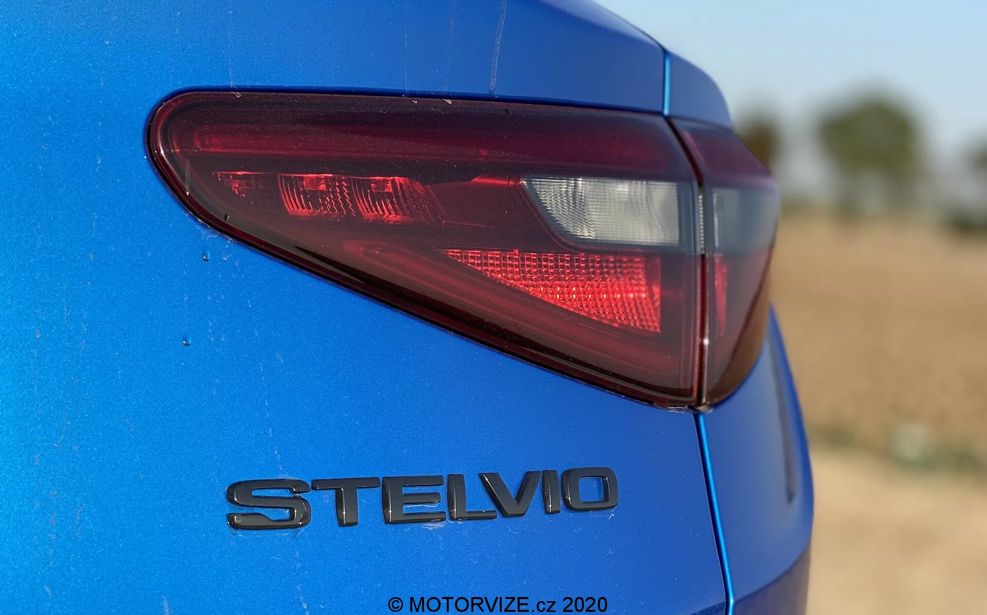 Detalhe em close-up da seção traseira do Alfa Romeo Stelvio 2017 azul, capturado de um ângulo baixo e ligeiramente fora de centro focando no lado traseiro esquerdo. Destaca a lanterna traseira moderna de LED e o emblema cromado 'STELVIO', mostrando o acabamento de pintura metálica distintiva. A pouca profundidade de campo mantém a lanterna traseira e o emblema em foco nítido contra o fundo desfocado. A luz natural do dia realça a profundidade da cor e os reflexos.