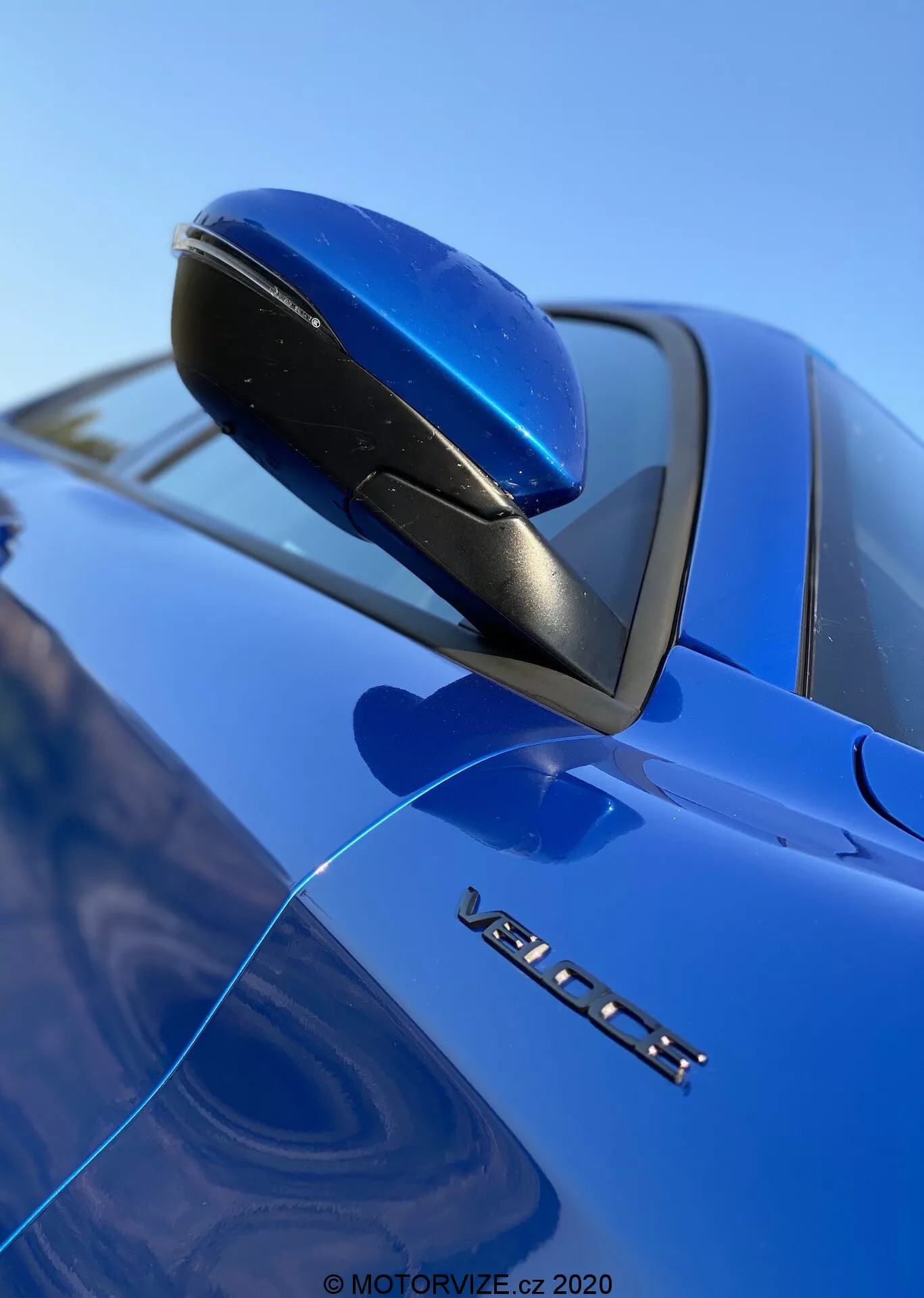 Vista aproximada e de ângulo baixo do espelho lateral de um Alfa Romeo Stelvio 2017 azul, enfatizando seu design aerodinâmico, base preta e indicador de seta integrado. A marca VOLVO e um emblema de modelo parcialmente visível estão abaixo do espelho, com foco nítido nesses detalhes contra um fundo desfocado de para-brisa e céu, destacando o acabamento da pintura brilhante do carro.