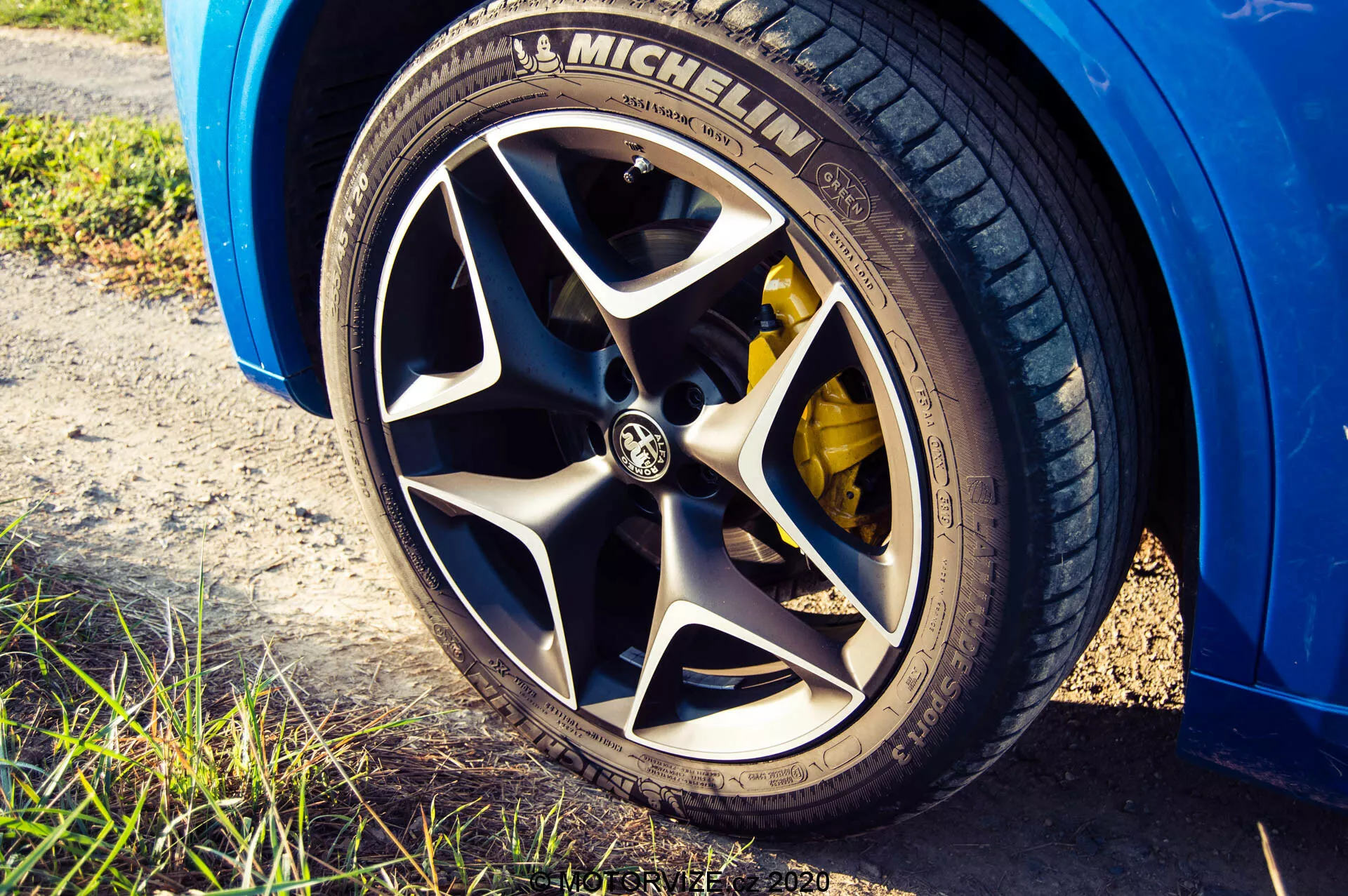 Imagem de perspectiva em close-up e ângulo baixo de uma roda do Alfa Romeo Stelvio 2017, exibindo um pneu Michelin com um padrão de piso visível, aro de liga leve com vários raios com logotipo da marca, pinça de freio de alto desempenho amarela, exterior do carro na cor azul brilhante, configurada em luz natural com um toque de ambiente ao ar livre e profundidade de campo rasa enfatizando a roda.
