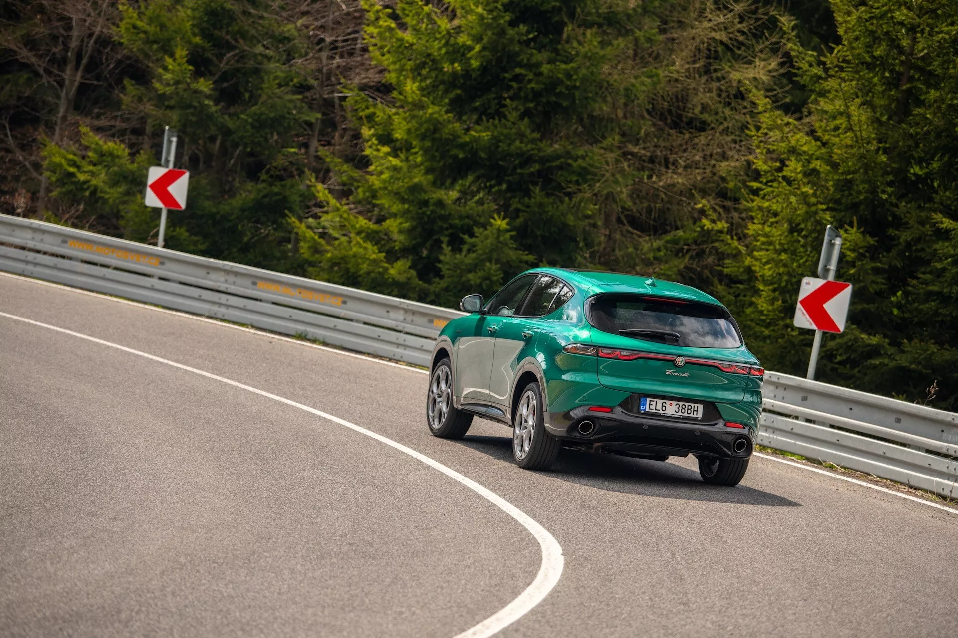 Alfa Romeo Tonale 2022 capturado de um ângulo traseiro de três quartos em uma estrada curva, destacando seu design esportivo hatchback, acabamento brilhante e características aerodinâmicas, incluindo um spoiler traseiro, rodas de liga leve e luzes traseiras LED.