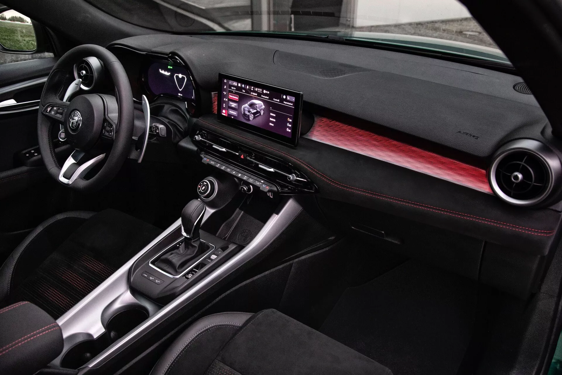 Interior do Alfa Romeo Tonale 2022 com um painel moderno, centro de infotenimento digital, detalhes em fibra de carbono e assentos com costuras vermelhas, visto da perspectiva do passageiro dianteiro