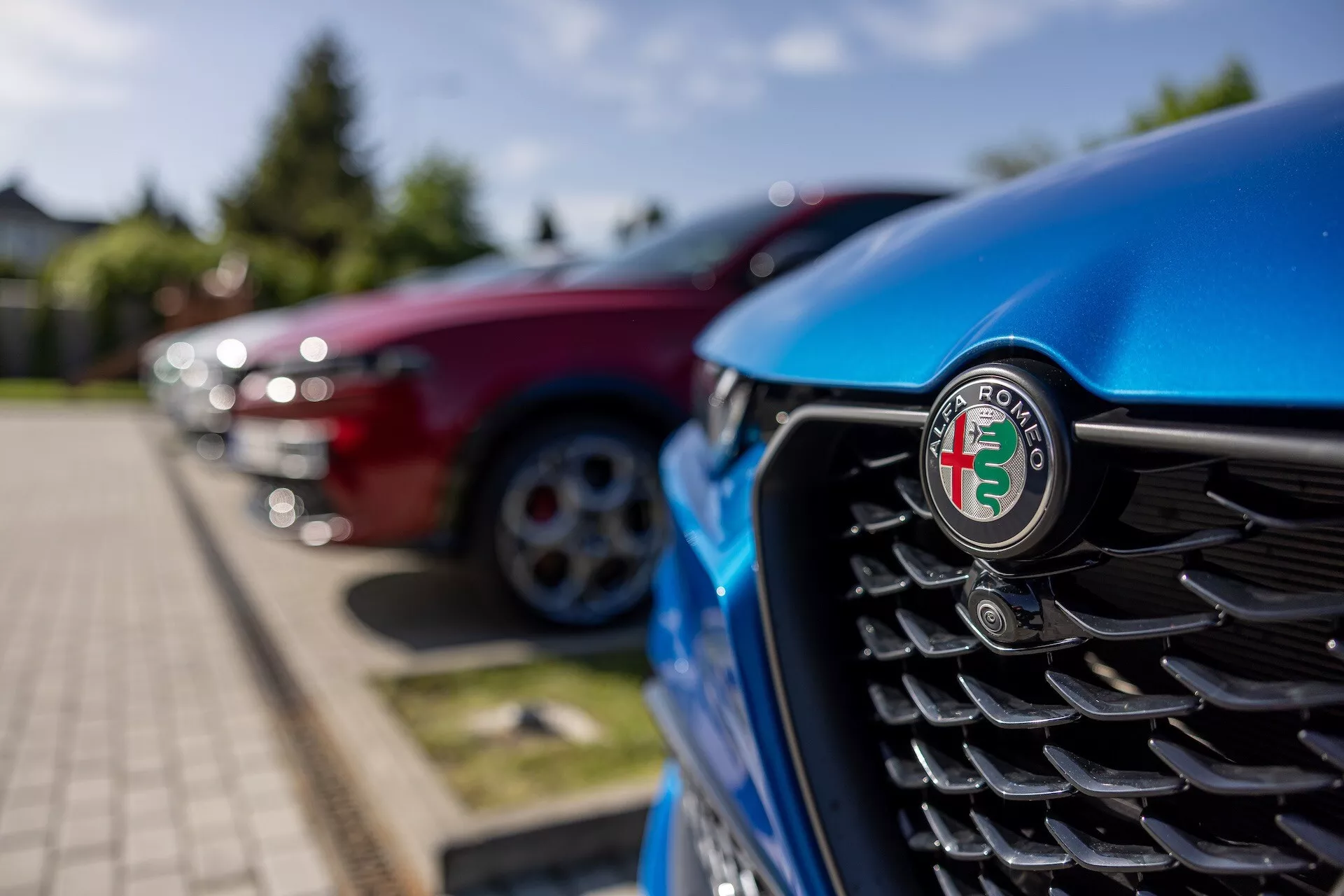 Vista detalhada da grelha frontal do Alfa Romeo Tonale 2022 azul, com foco no emblema, apresentando lâminas verticais e um fundo desfocado de outros veículos