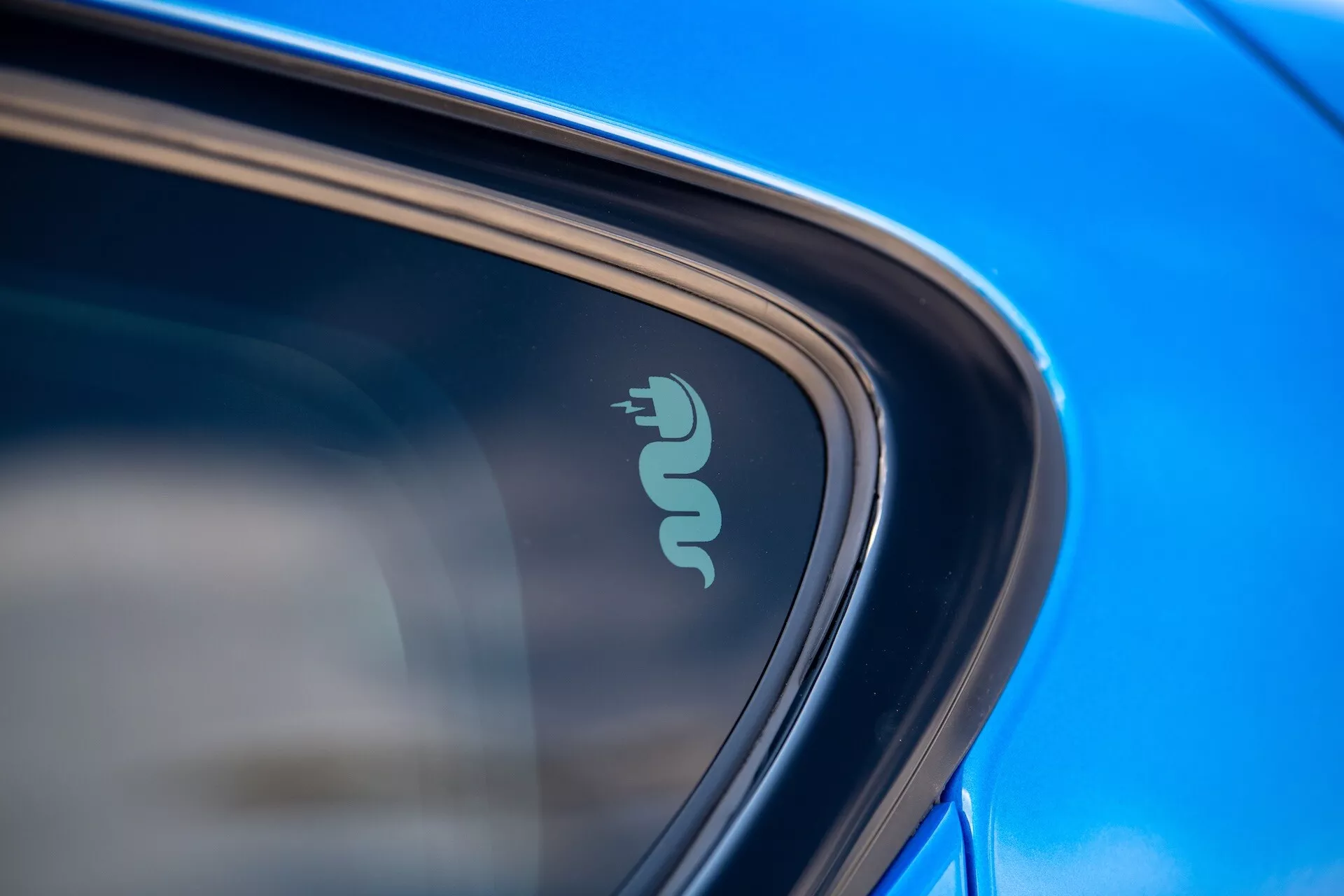 Close-up detalhado do Alfa Romeo Tonale 2022 exibindo distintivo tecnológico indicativo de capacidade elétrica ou híbrida perto da janela do carro, realçado por pintura azul reflexiva e design elegante à luz natural do dia