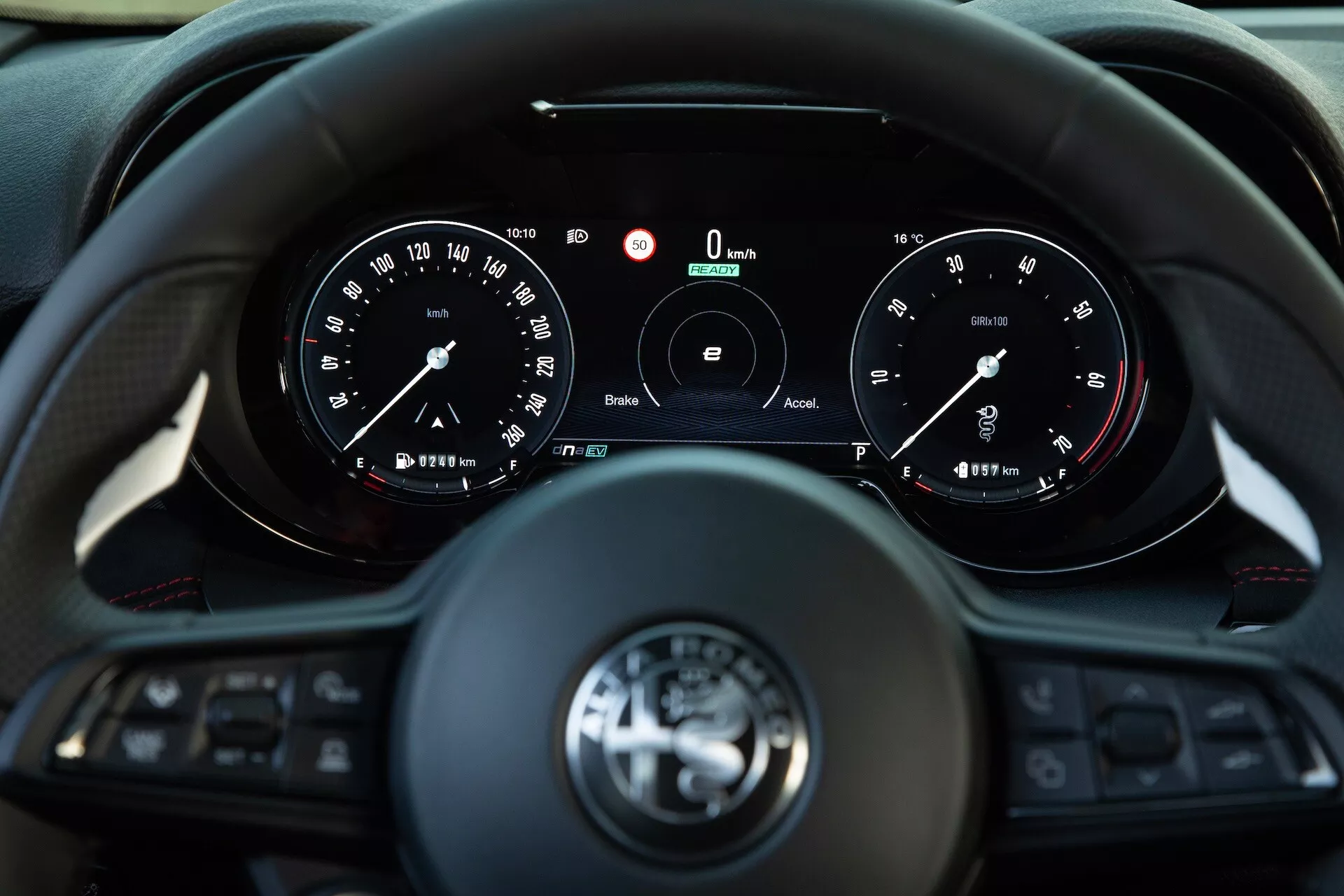 Interior do Alfa Romeo Tonale 2022 mostrando a visão do condutor de um volante multifuncional com três raios e um conjunto de instrumentos com mostradores analógicos e um display digital central indicando velocidade e RPM zero, apresentando tecnologias de conectividade e assistência ao condutor