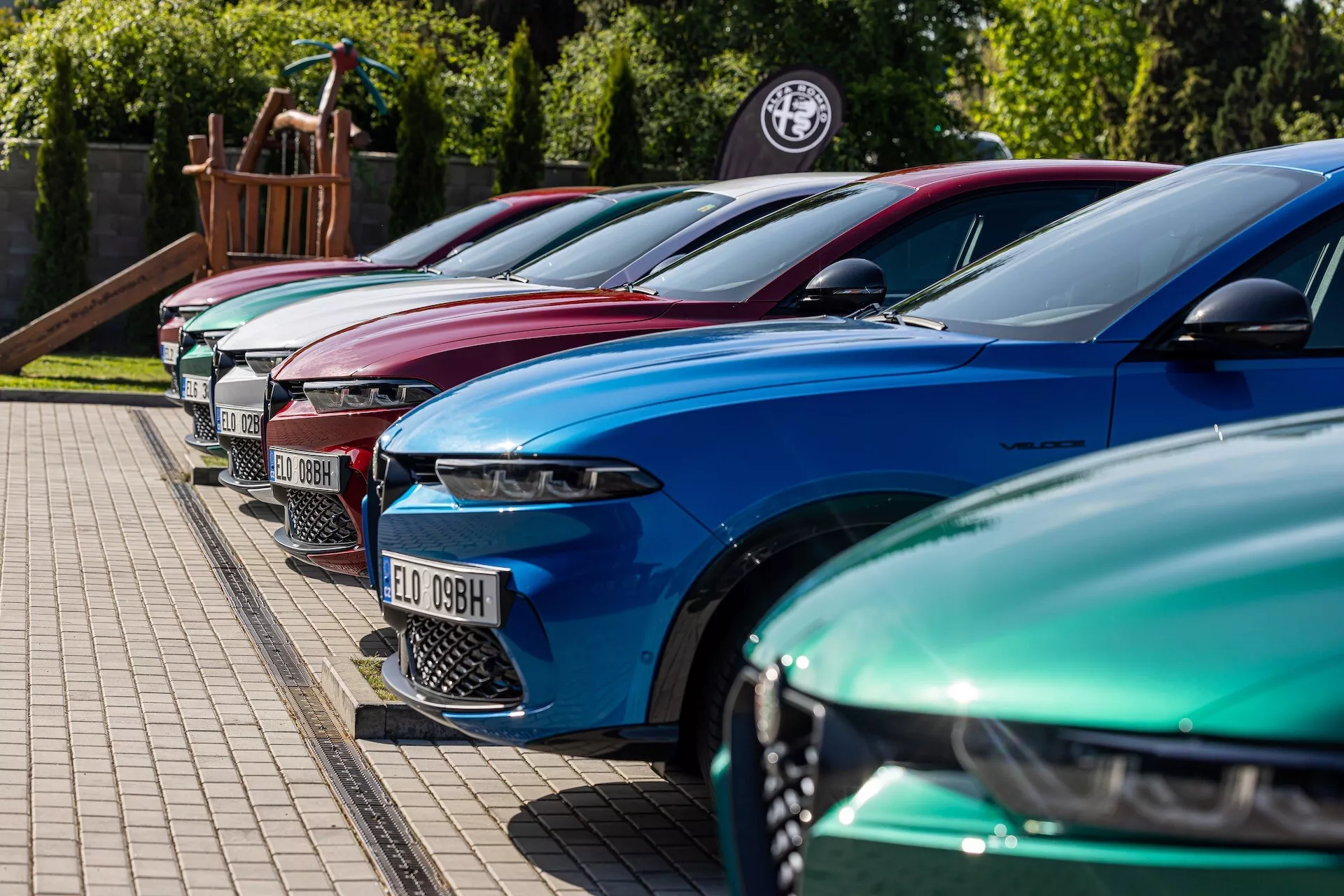 Linha Alfa-Romeo Tonale 2022 em cores vibrantes mostrando design de luxo com faróis LED avançados, estacionados em formação diagonal com foco no lado frontal esquerdo de um ângulo de altura baixa a média em uma área pavimentada ao lado de uma borda gramada, possivelmente em uma concessionária ou evento de entusiastas de carro
