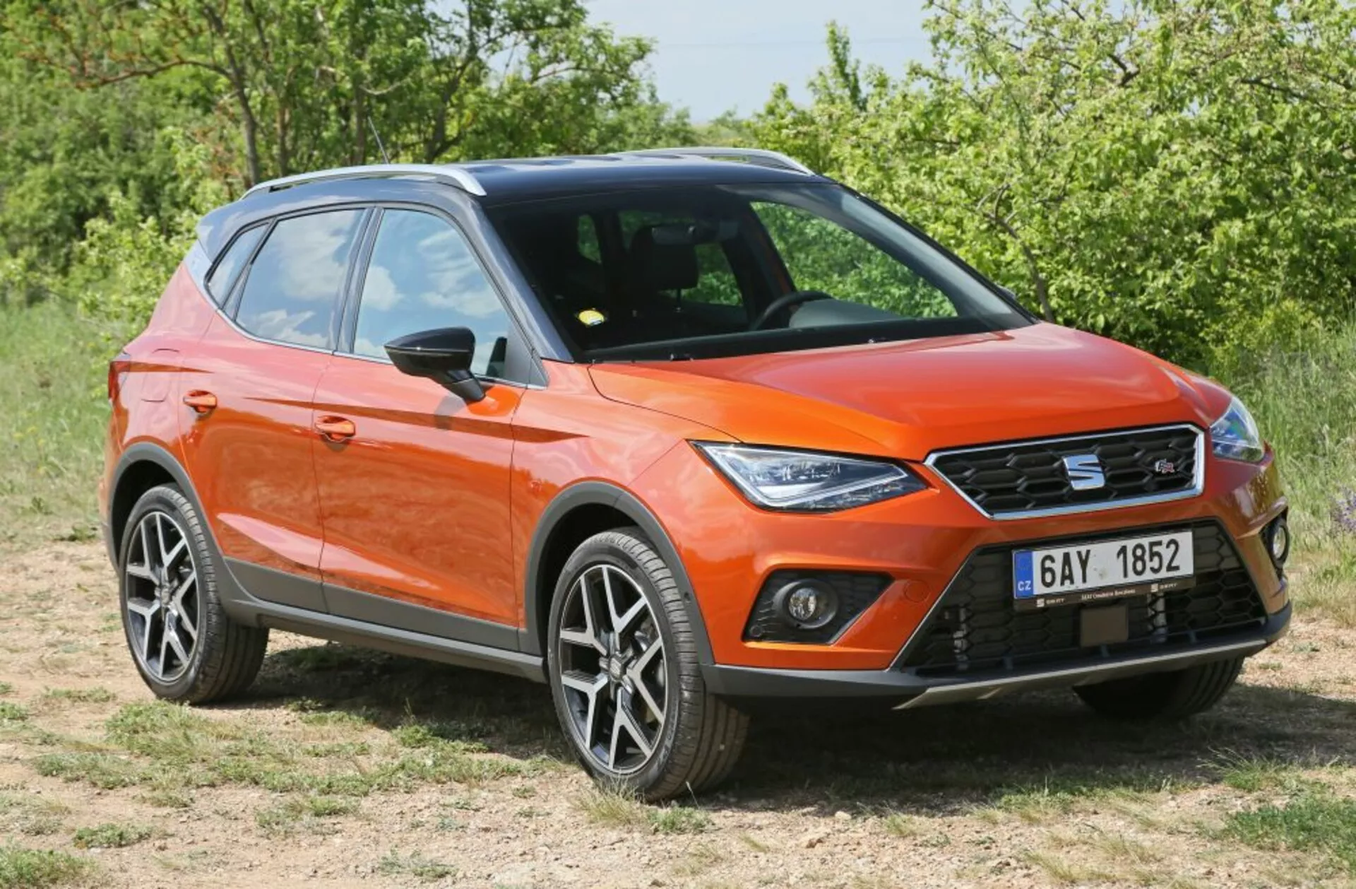 Crossover/SUV laranja estacionado na grama em visão frontal de três quartos mostrando luzes diurnas LED, design moderno e afiado com rodas de liga leve e barras de teto sob a luz do sol