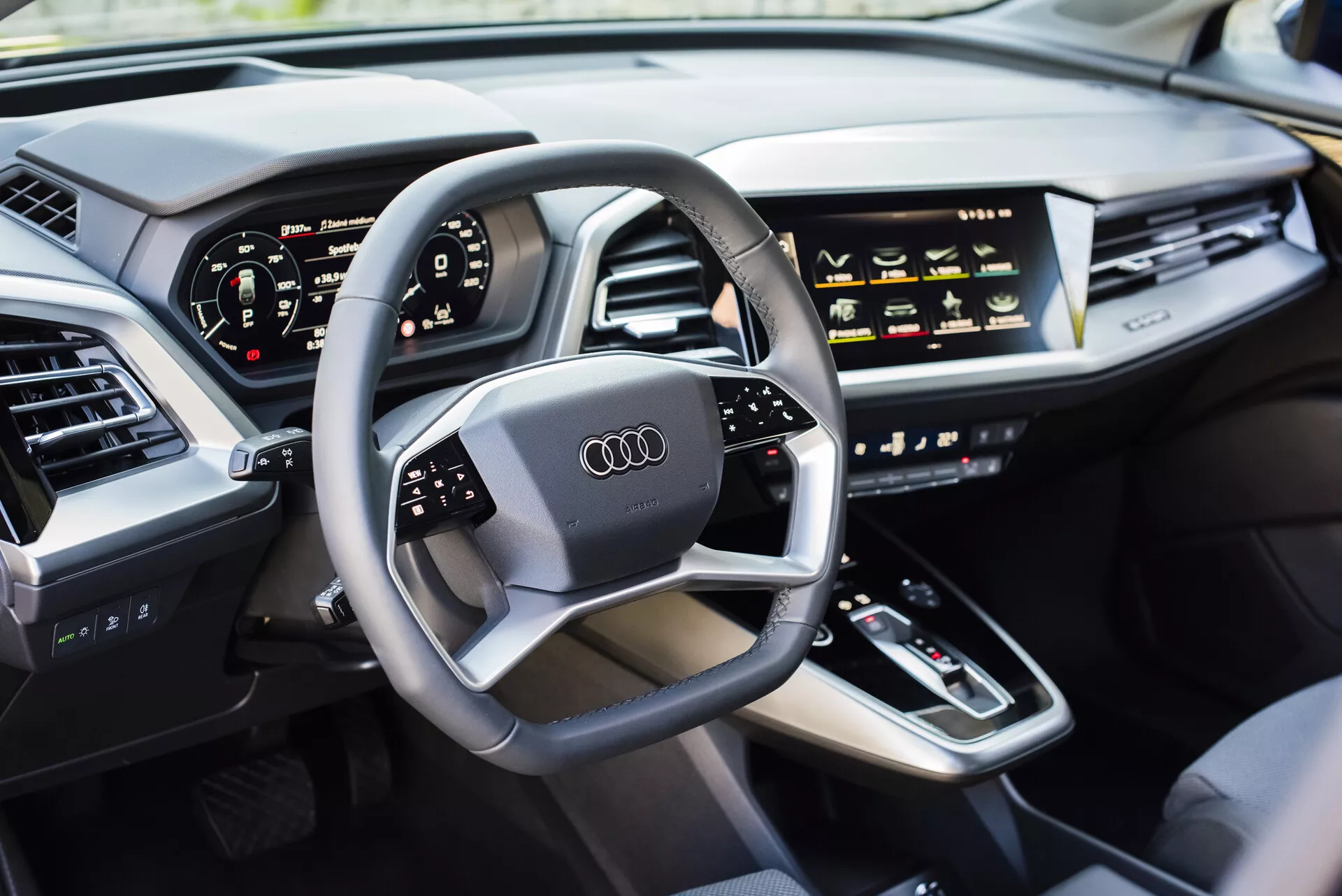 Vista interior de um Audi Q4 e-tron 2021 pelo lado do motorista, destacando o volante multifuncional com logotipo da Audi, cluster de instrumentos digital, console central com alavanca de câmbio, controles sensíveis ao toque HVAC, display de infotainment, painel elegante com saídas de ar integradas, e uma porta acolchoada de couro de luxo, exibindo tecnologia automotiva moderna e características de veículos de luxo.