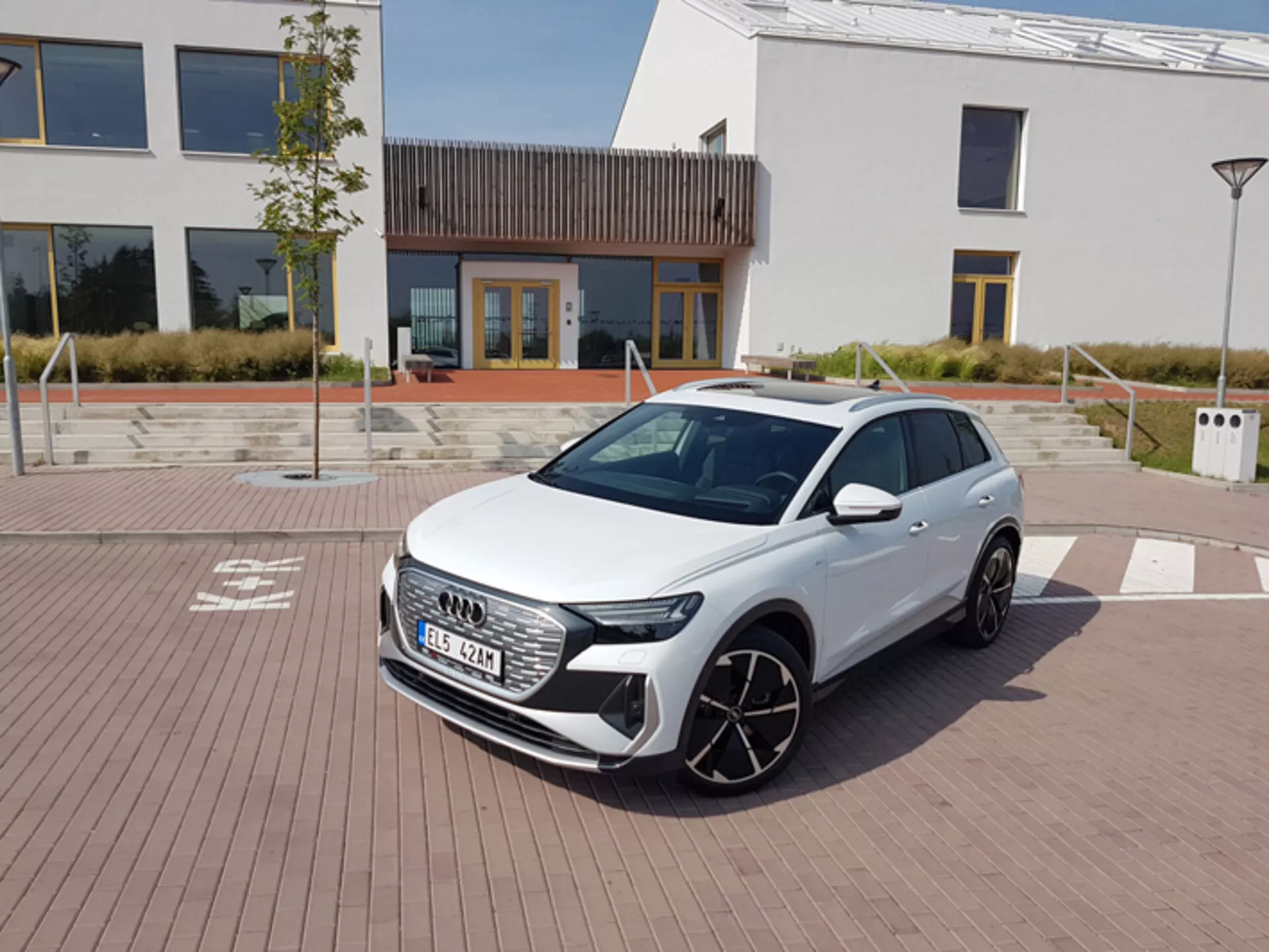 Audi Q4 e-tron 2021 branco, exibindo seus modernos faróis de LED, grade frontal hexagonal e grandes rodas de liga leve, estacionado em uma área pavimentada com padrão de tijolos com arquitetura urbana e uma estação de carregamento ao fundo, sob um céu claro ao meio-dia.