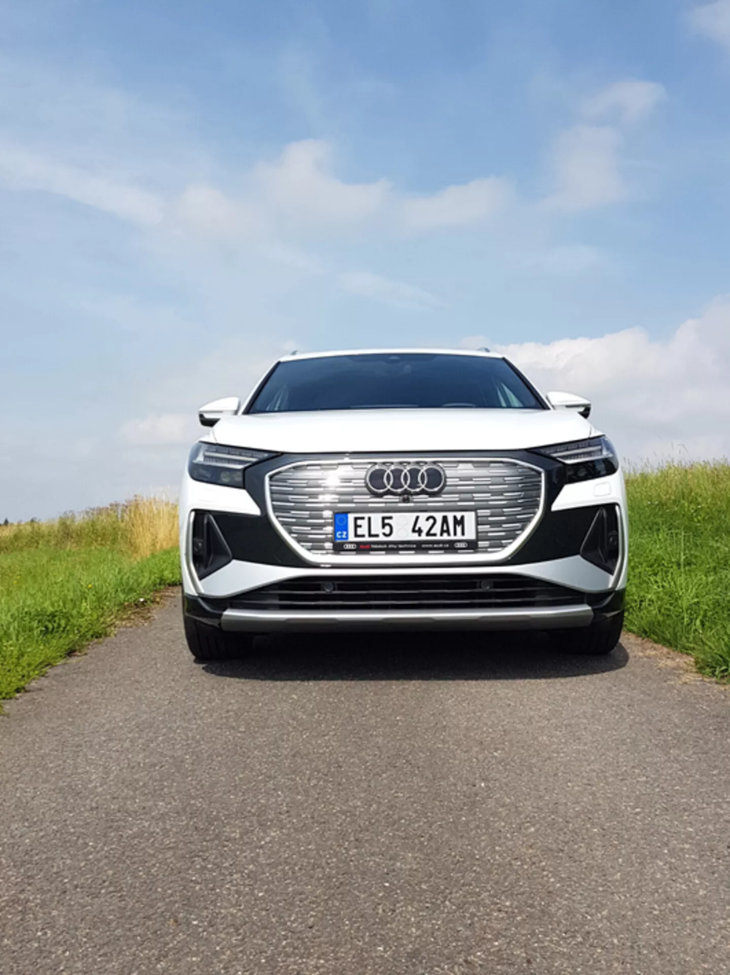 Vista frontal centrada do Audi Q4 e-tron 2021 mostrando grande grade hexagonal com emblema Audi, faróis LED, placa de matrícula europeia, possíveis sensores ADAS, entradas de ar, pintura metálica e acabamento cromado sob céu parcialmente nublado em uma estrada de asfalto com lados gramados.
