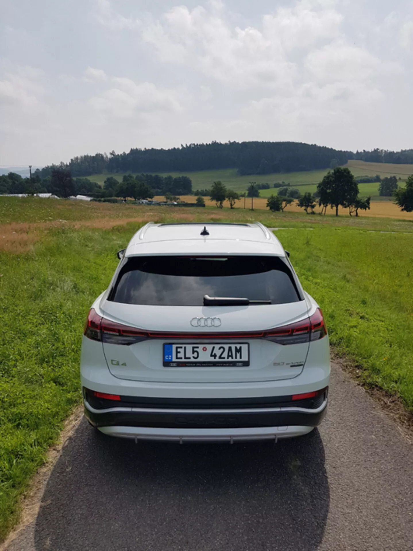 Vista traseira de um Audi Q4 e-tron 2021 prateado em uma estrada pavimentada estreita no meio de uma paisagem pastoril com campos e árvores sob um céu parcialmente nublado, destacando seu design elegante, barra de luz de freio, spoiler traseiro, sensores de estacionamento traseiro e rodas de liga leve bicolores.