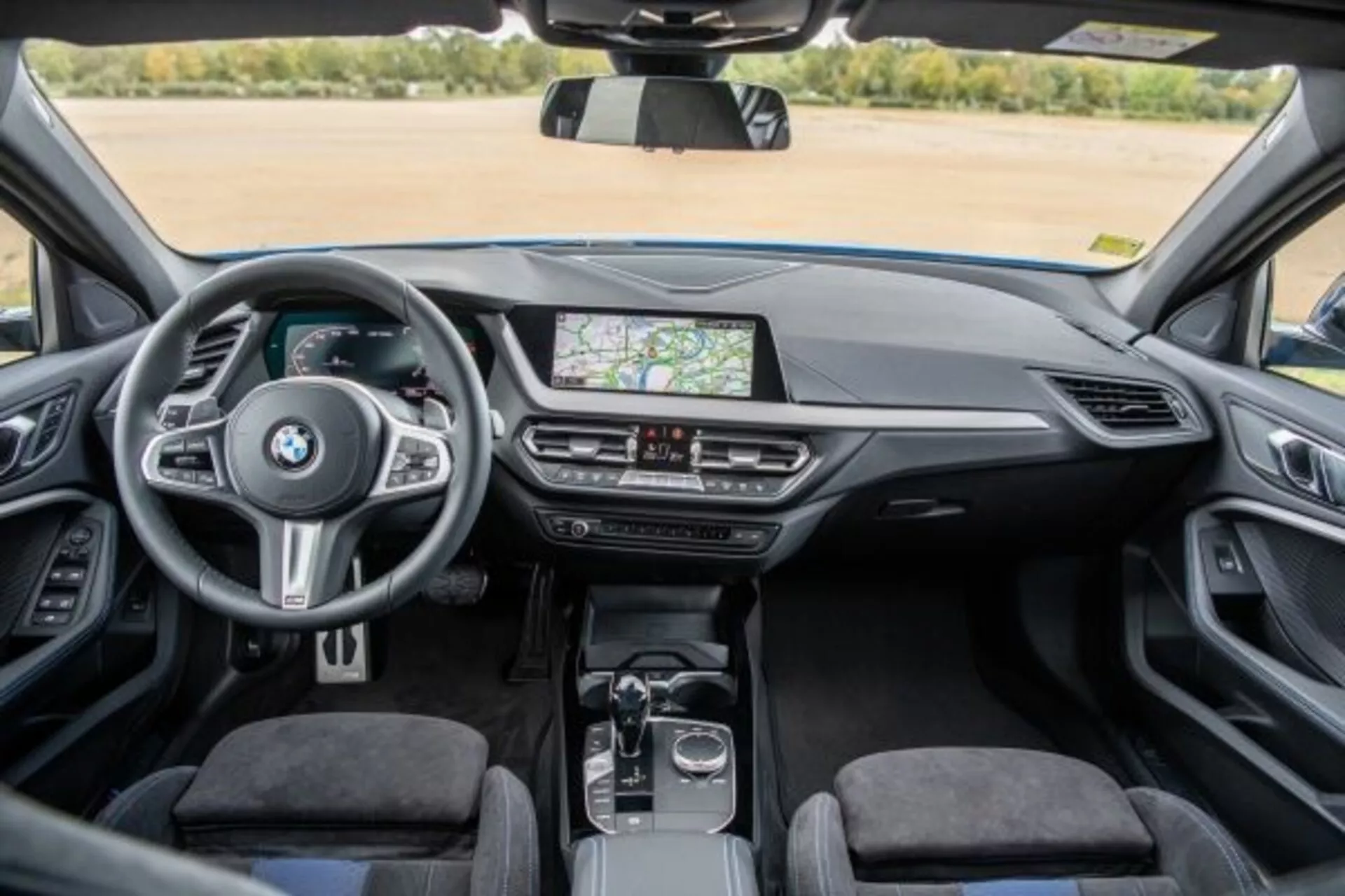 Vista interior do BMW Série 1 M Performance 2019 a partir do assento traseiro mostrando o painel, console central e volante com controles montados, sistema de infotenimento com tela sensível ao toque exibindo navegação GPS, ventilações, controles do clima, alavanca de câmbio automática, disco rotativo e touchpad no console central, estofamento premium e uma cabine clara e bem iluminada
