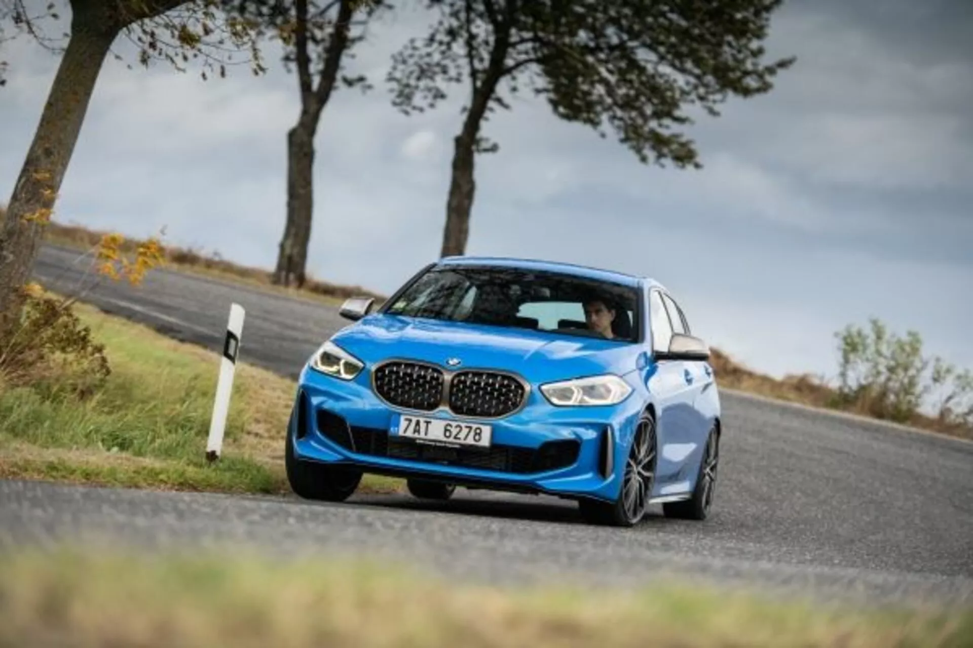 Fotografia dinâmica de baixo ângulo de um moderno sedã esportivo azul BMW Série 1 M Performance 2019 em movimento em uma estrada rural, exibindo seu design, a grade característica em forma de rim, faróis LED agressivos, para-choque dianteiro esculpido com entradas de ar e rodas de liga leve