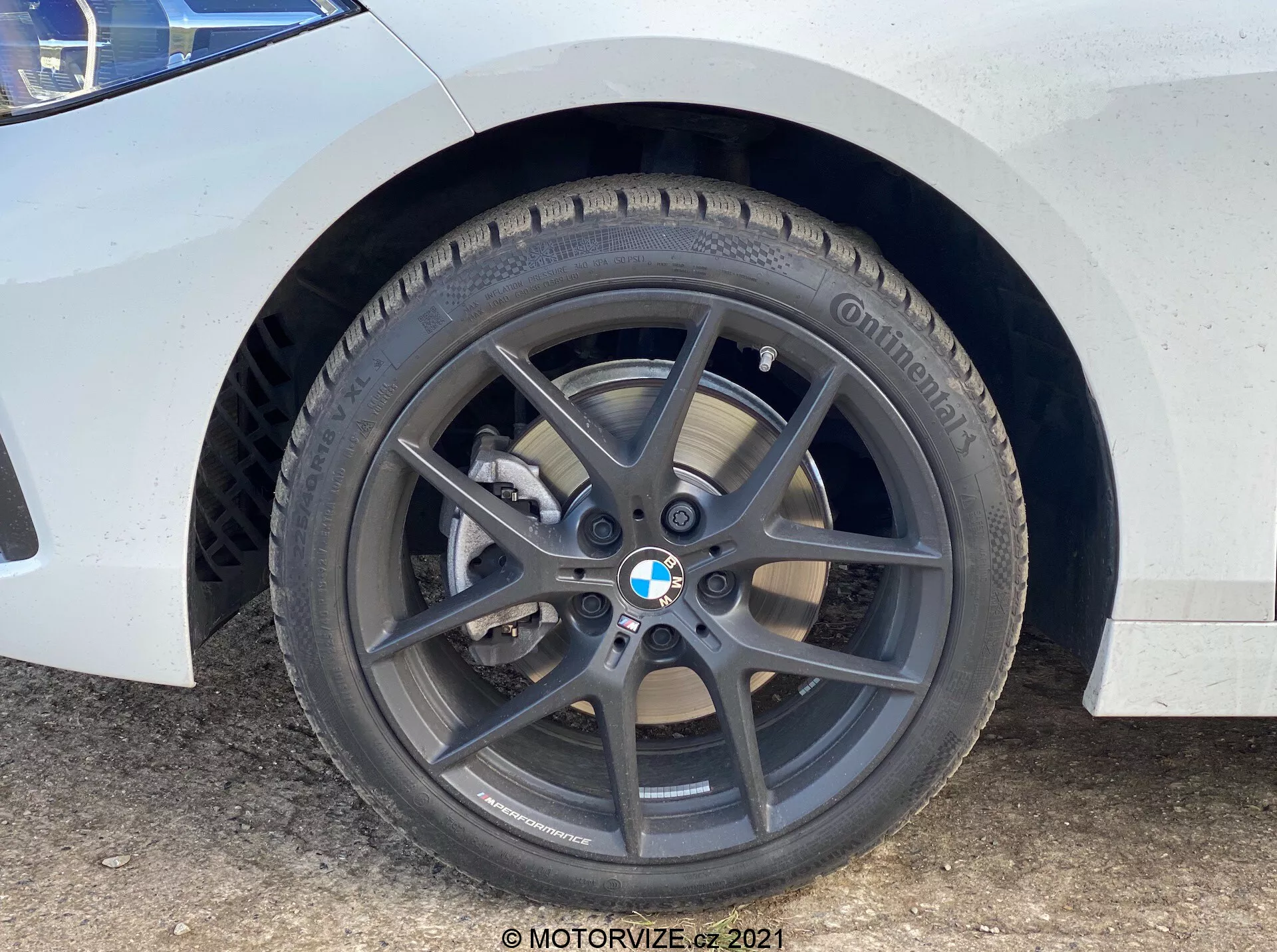 Close-up da roda dianteira e para-lama do BMW Série 1 2019 (BMW-116i). Perspectiva ao nível do solo destaca a roda de liga leve de cinco raios cinza escuro com emblema BMW, pneu Continental com banda de rodagem visível, disco de freio ventilado e pinça de freio de pistão único prateada. A pintura branca com manchas de sujeira e água indica um estado de uso.