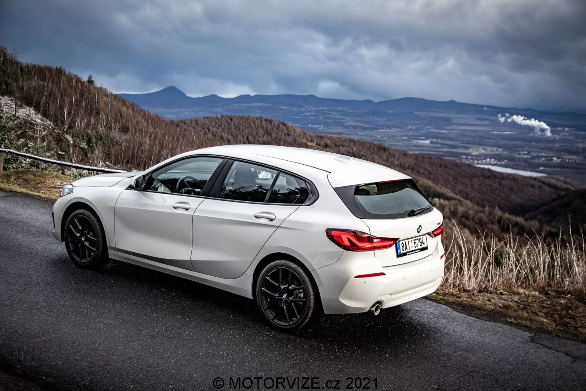 Vista lateral traseira angulada de um hatchback BMW 116i branco de 2019 em uma estrada de asfalto com um céu dramático e nublado. A imagem destaca o design traseiro do veículo, os arcos das rodas traseiras, as rodas de liga leve de cor escura, o sistema de escape duplo e o perfil lateral, incluindo as portas traseira do passageiro e do motorista. O cenário sugere uma estrada em uma colina ou montanha, enfatizando os atributos esportivos e dinâmicos do carro. Marca d'água do fotógrafo 'MOTORWIZE.cz 2021' presente.