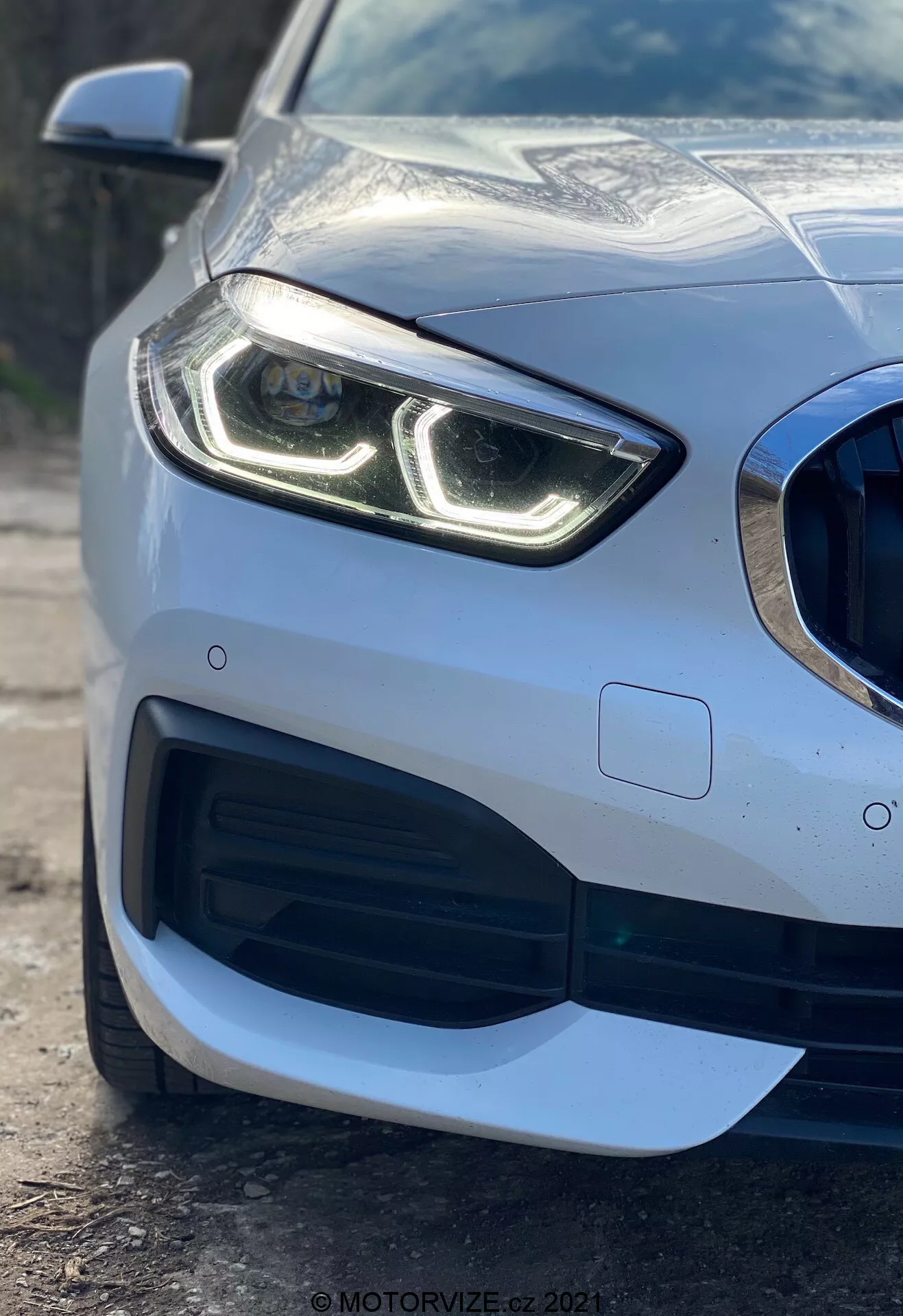 Vista de perto da seção frontal esquerda de um carro BMW Série 1 2019 (BMW 116i) destacando seu farol moderno com luzes diurnas em LED distintivas, grade com padrão de favo de mel e grande entrada de ar, design aerodinâmico sofisticado no para-choque dianteiro e sensor de estacionamento embutido no para-choque, mostrando o acabamento de pintura metálica premium e a carroceria refletiva.