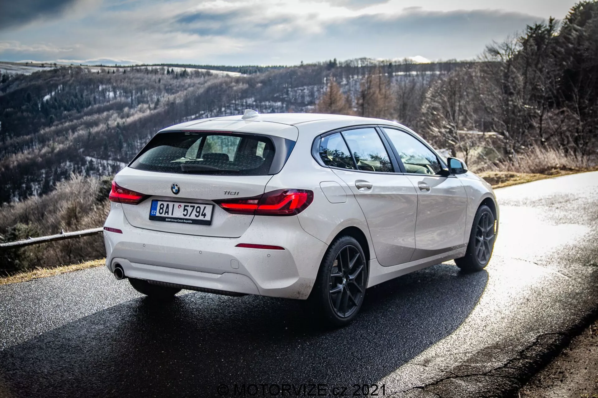 Uma vista traseira três quartos dinâmica de um BMW 116i 2019 branco, mostrando seu lado direito e traseiro sob a luz do dia com raios de sol claros e sombras. A imagem enfatiza os detalhes do design, incluindo realces pretos no difusor traseiro, aros das rodas e capas dos espelhos laterais, complementados por rodas de liga leve e um sistema de escape duplo, sugerindo uma variante mais esportiva. A estrada curva para a direita com um fundo montanhoso ou colinoso, ideal para entusiastas de condução.