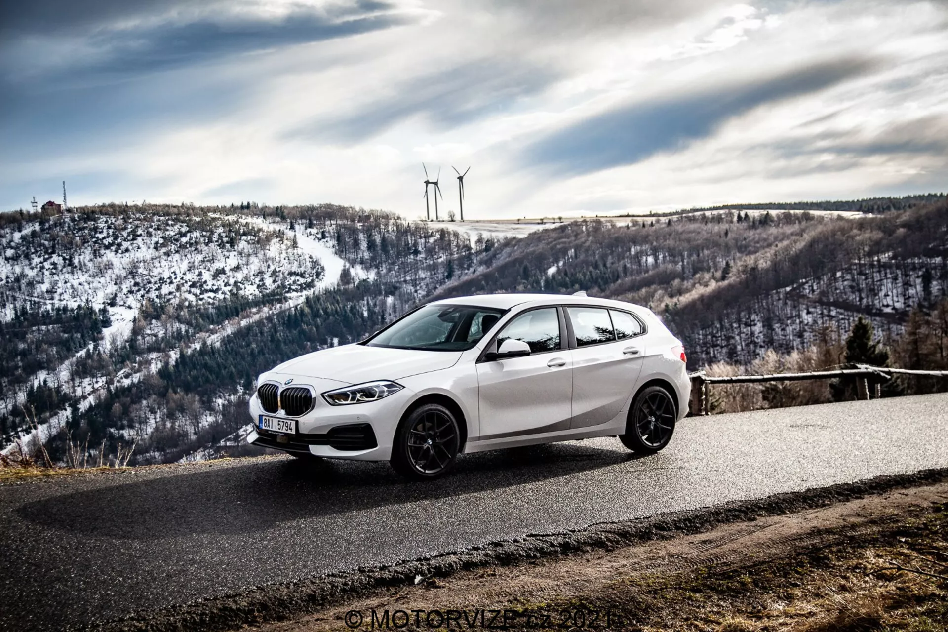 Um carro hatchback BMW branco, possivelmente um BMW 1er-Reihe 2019 (BMW 116i), está estacionado em uma estrada de asfalto curva cercada por uma paisagem montanhosa e nevada com turbinas eólicas. A foto, tirada de um ângulo frontal lateral, destaca a grade dianteira, faróis, linhas aerodinâmicas e rodas de liga leve do carro sob um céu nublado.