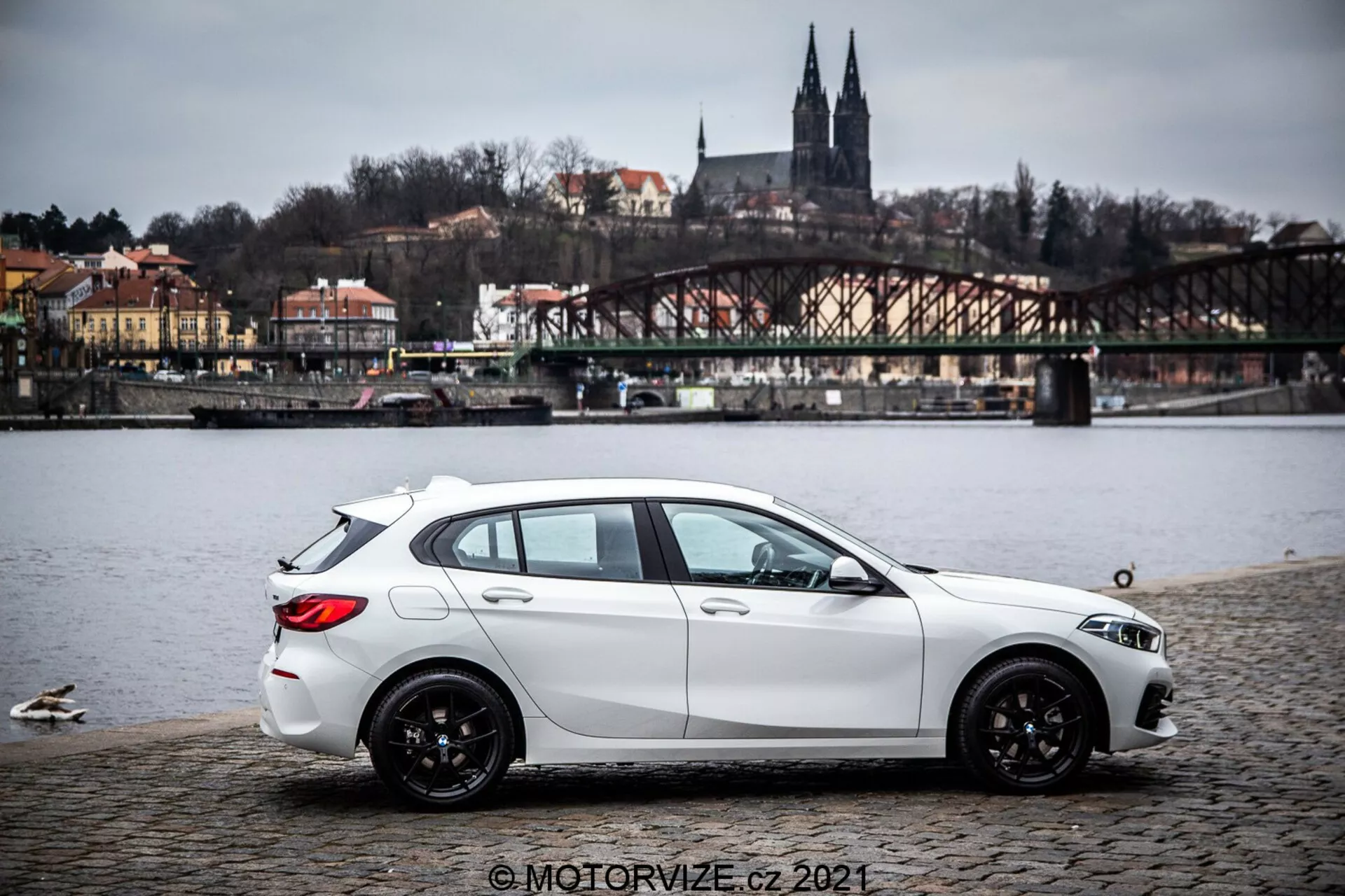 Um hatchback BMW Série 1 de 2019 (BMW-116i) branco, estacionado em uma superfície de paralelepípedos perto de um corpo d'água com patos, mostrando um perfil lateral com uma leve vista traseira. O carro possui linhas esculpidas dinâmicas, linha de teto inclinada, rodas de liga leve pretas com múltiplos raios e faróis e espelhos laterais visíveis. Ao fundo está uma ponte, edifícios em uma colina e elementos arquitetônicos que sugerem um ambiente europeu. A iluminação é difusa sob um céu nublado e a imagem inclui uma marca d'água © MOTORVIZE.cz 2021.