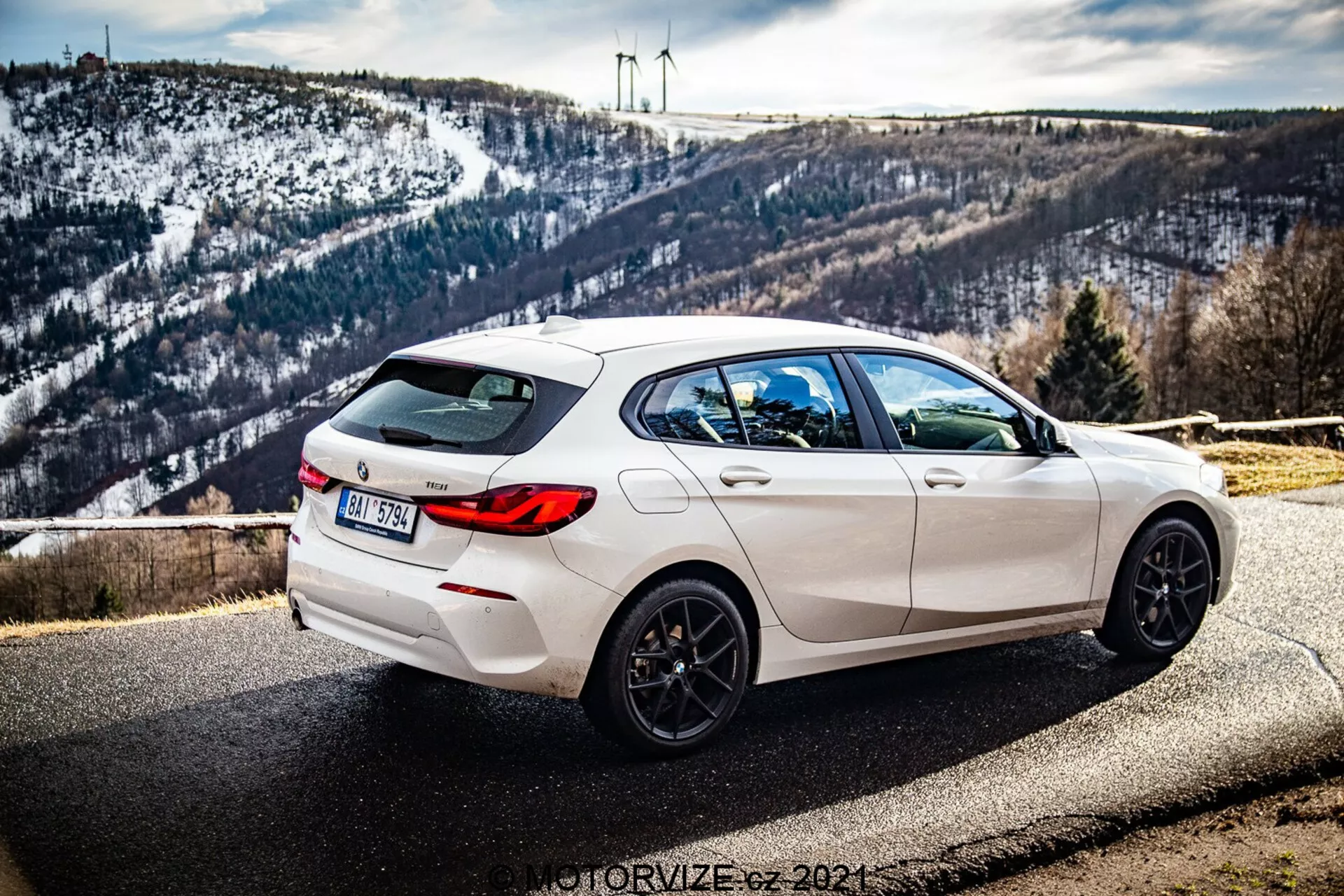 A imagem mostra um BMW Série 1 (BMW-116i) de 2019 branco estacionado à beira da estrada com um cenário pitoresco ao fundo, incluindo uma paisagem montanhosa coberta de neve e turbinas eólicas no horizonte. A fotografia é tirada de um ângulo traseiro de três quartos, exibindo claramente o lado direito do carro. A vista destaca as linhas de design elegantes, o spoiler traseiro, as rodas de liga leve pretas de cinco raios, as saídas duplas de escape e os vidros traseiros escurecidos. O carro também possui limpador de vidro traseiro e uma antena tipo barbatana de tubarão integrada no teto. A profundidade de campo mantém o carro em foco agudo com luz natural destacando sua estética esportiva enquanto o fundo permanece sutilmente detalhado.