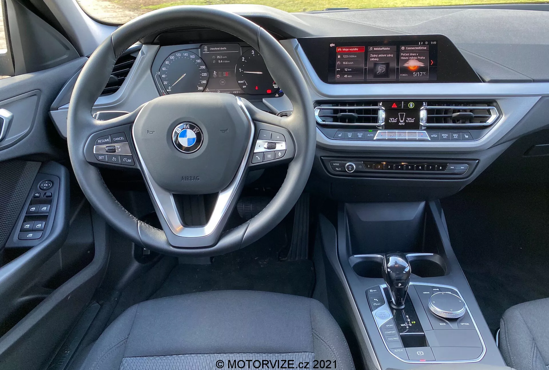 Vista interior do BMW Série 1 (BMW 116i) 2019 da perspectiva do assento do passageiro dianteiro, mostrando o volante multifuncional com controles montados, painel de instrumentos digital, grande tela sensível ao toque do sistema de infoentretenimento, sistema de controle climático, alavanca de câmbio automática, seletor de modo de condução e o botão de controle iDrive.