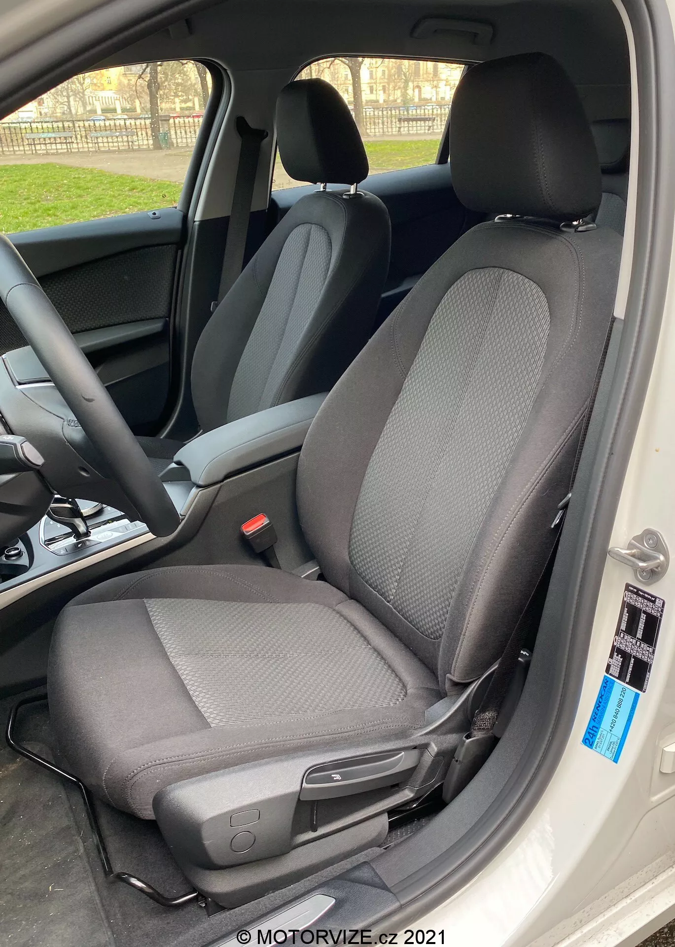 Close-up do interior do BMW 1er-Reihe 2019 (BMW-116i) focando no assento do motorista com um design de tecido moderno de duas cores, predominantemente preto com padrões em cinza claro, tirada de um ângulo lateral com a porta do carro totalmente aberta. O assento possui um encosto de cabeça contra a luz, criando um efeito de silhueta ao redor das bordas, e inclui uma alavanca para ajustes manuais para frente ou para trás, bem como um pequeno botão preto próximo à borda do interior do carro. A luz natural do dia realça o foco nítido no assento, com um fundo desfocado mostrando o volante e o painel.