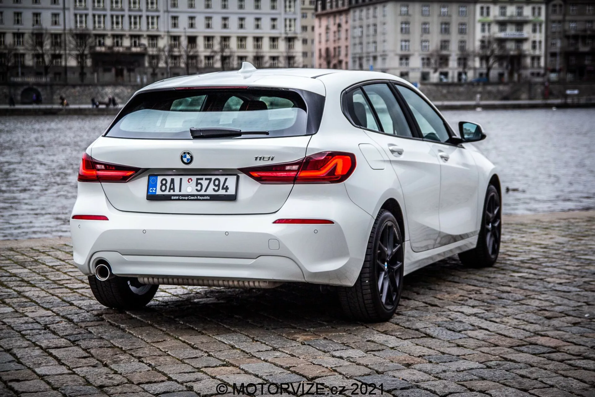 BMW Série 1 de 2019 (116i) hatchback branco, mostrando vista de três quartos traseira com elementos detalhados, incluindo luzes traseiras, tampa do porta-malas, para-choque traseiro, tubos de escape e parte das rodas traseiras, estacionado em uma superfície de paralelepípedo molhada perto de um corpo d'água. A imagem destaca o spoiler traseiro esportivo, clusters de luzes traseiras modernas e emblemas visíveis da BMW e do nível de acabamento.