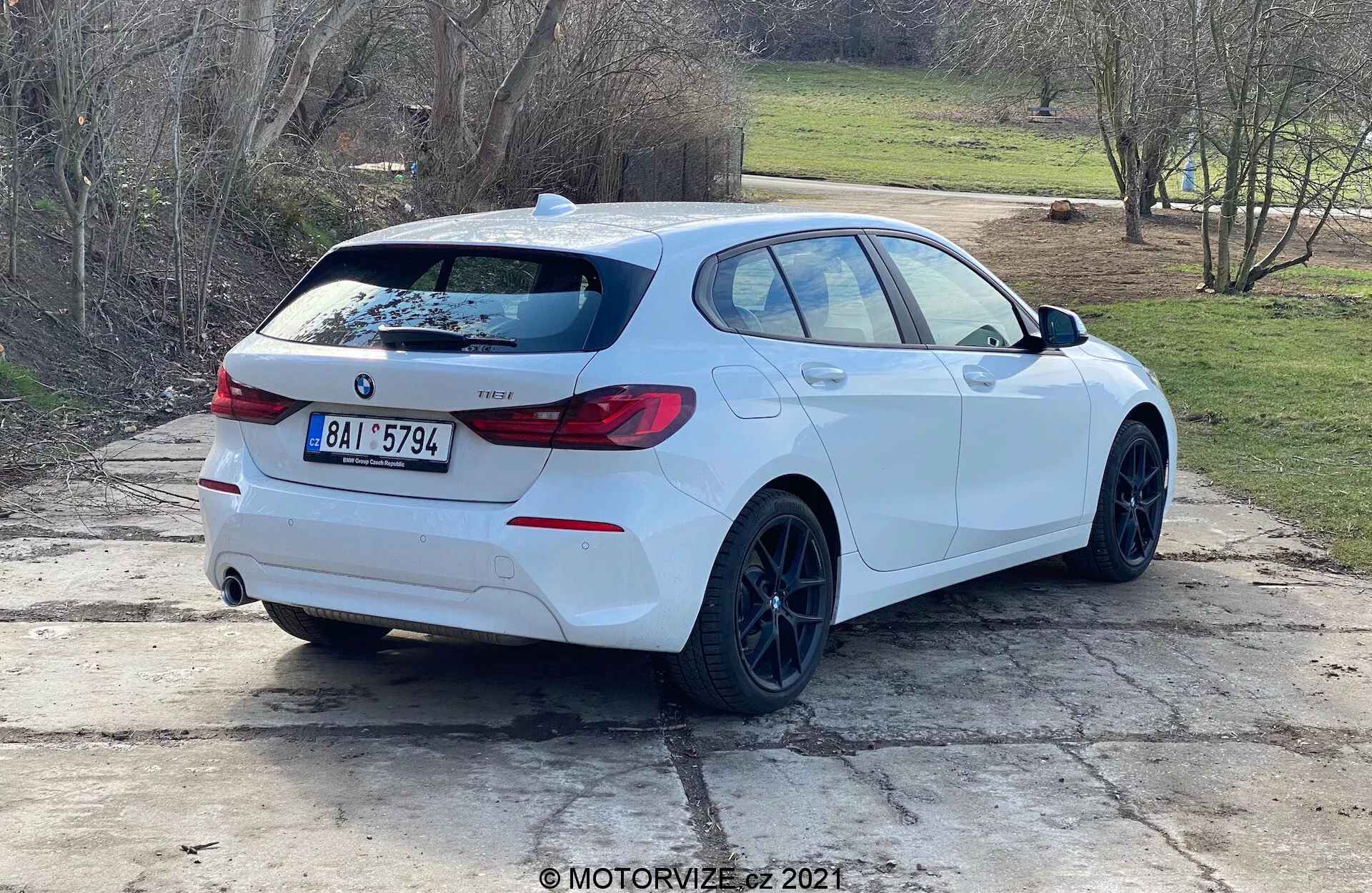Vista traseira de três quartos de um hatchback BMW Série 1 branco (modelo BMW 116i 2019) estacionado ao ar livre durante o dia. O perfil lateral e a parte traseira do carro são claramente visíveis, com o emblema BMW, designação do modelo, luzes traseiras distintas, spoiler traseiro, jantes pretas, estilo de carroçaria hatchback, tubos de escape duplos, limpa-vidros traseiro, vidros ligeiramente escurecidos e retrovisores com indicadores de sinal de mudança de direção integrados. O carro está posicionado em um espaço de concreto com manchas de grama e um cenário rural ou suburbano ao fundo.