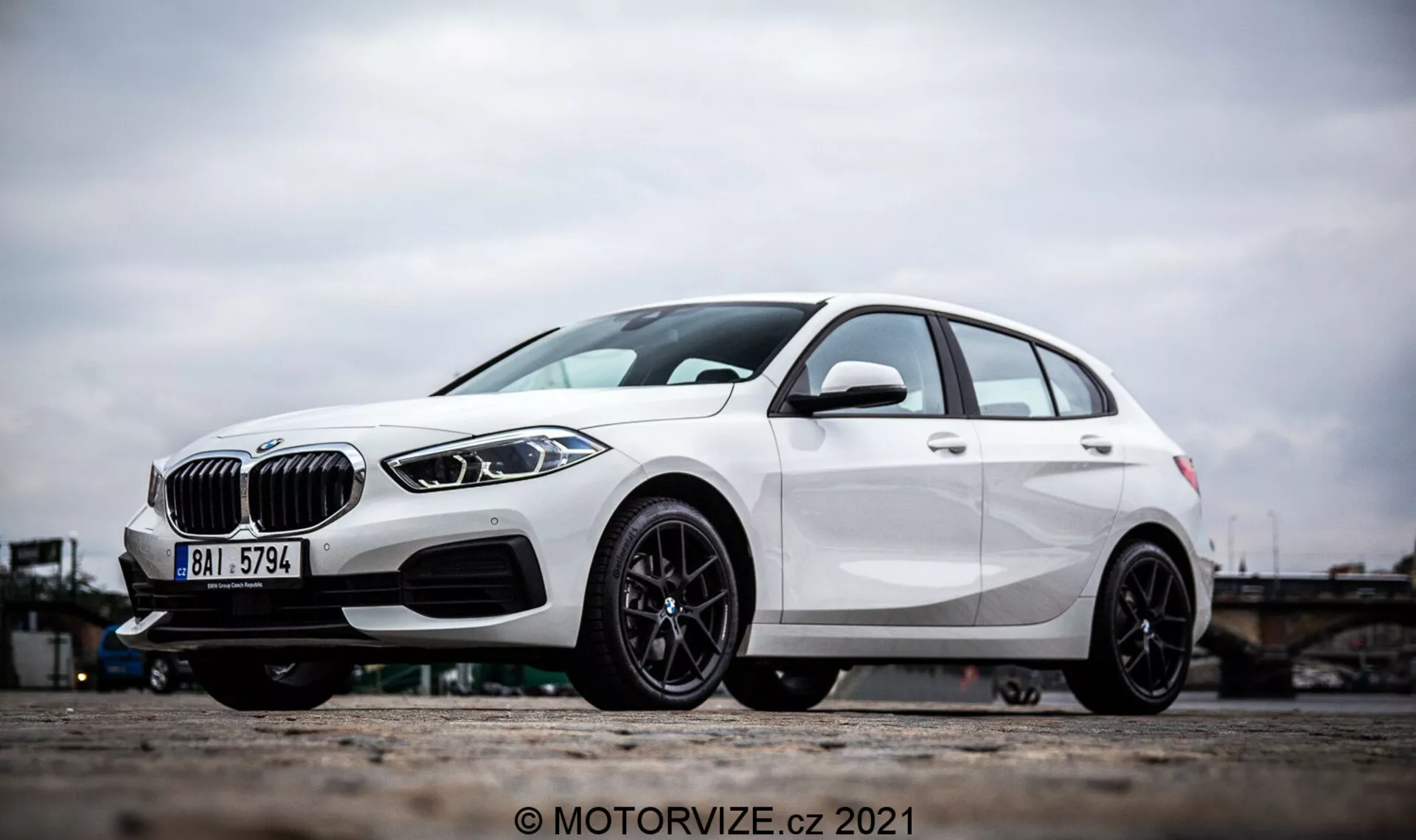 Vista lateral angular de um BMW Série 1 2019 (BMW 116i) branco, com elementos de design proeminentes como a grade em forma de rim, faróis de LED elegantes e para-choque dianteiro com entradas de ar e possíveis sensores de radar para assistência ao motorista. A foto se concentra nas rodas de liga leve e na parte inferior do veículo, destacando o design esportivo de várias raias pretas e o freio a disco de alto desempenho. As linhas de caráter e a escultura aerodinâmica do hatchback enfatizam ainda mais seu exterior moderno e dinâmico.