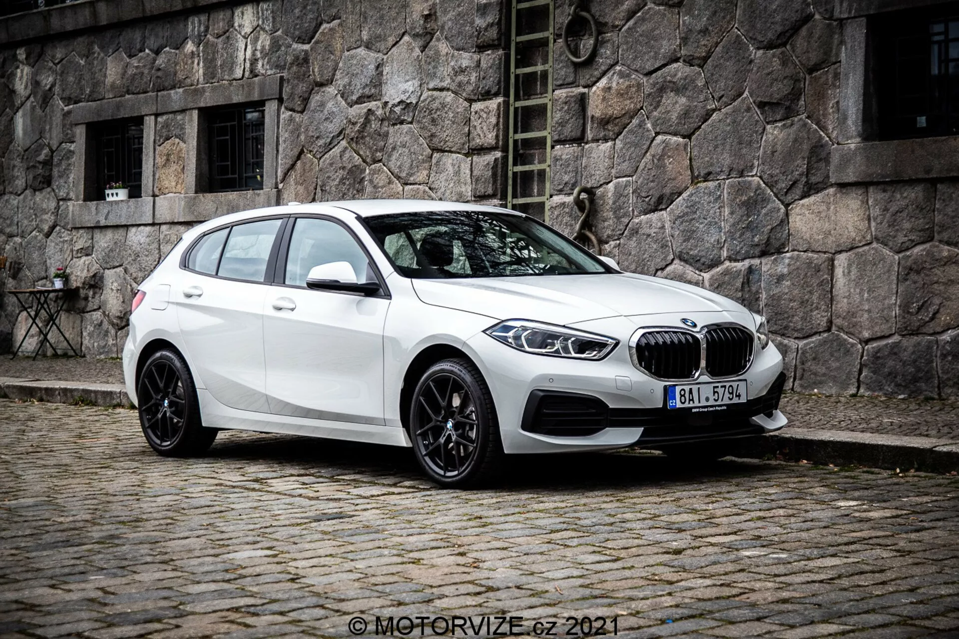Um BMW 1er-Reihe 2019 (BMW 116i) hatchback branco visto a partir de um ângulo frontal de três quartos, mostrando o lado esquerdo do veículo. A foto, tirada de um ângulo ligeiramente inferior, acentua a postura e as linhas de design do carro. Está posicionado numa rua de paralelepípedos com um antigo prédio de pedra ao fundo, criando uma cena urbana clássica. A grade em forma de rim e o emblema redondo BMW são visíveis na frente, ladeados por elegantes faróis LED angulares. O carro tem rodas de liga leve pretas com vários raios, uma linha de teto inclinada, espelhos laterais na cor do corpo com indicadores de direção integrados e vidros escurecidos para os passageiros traseiros.