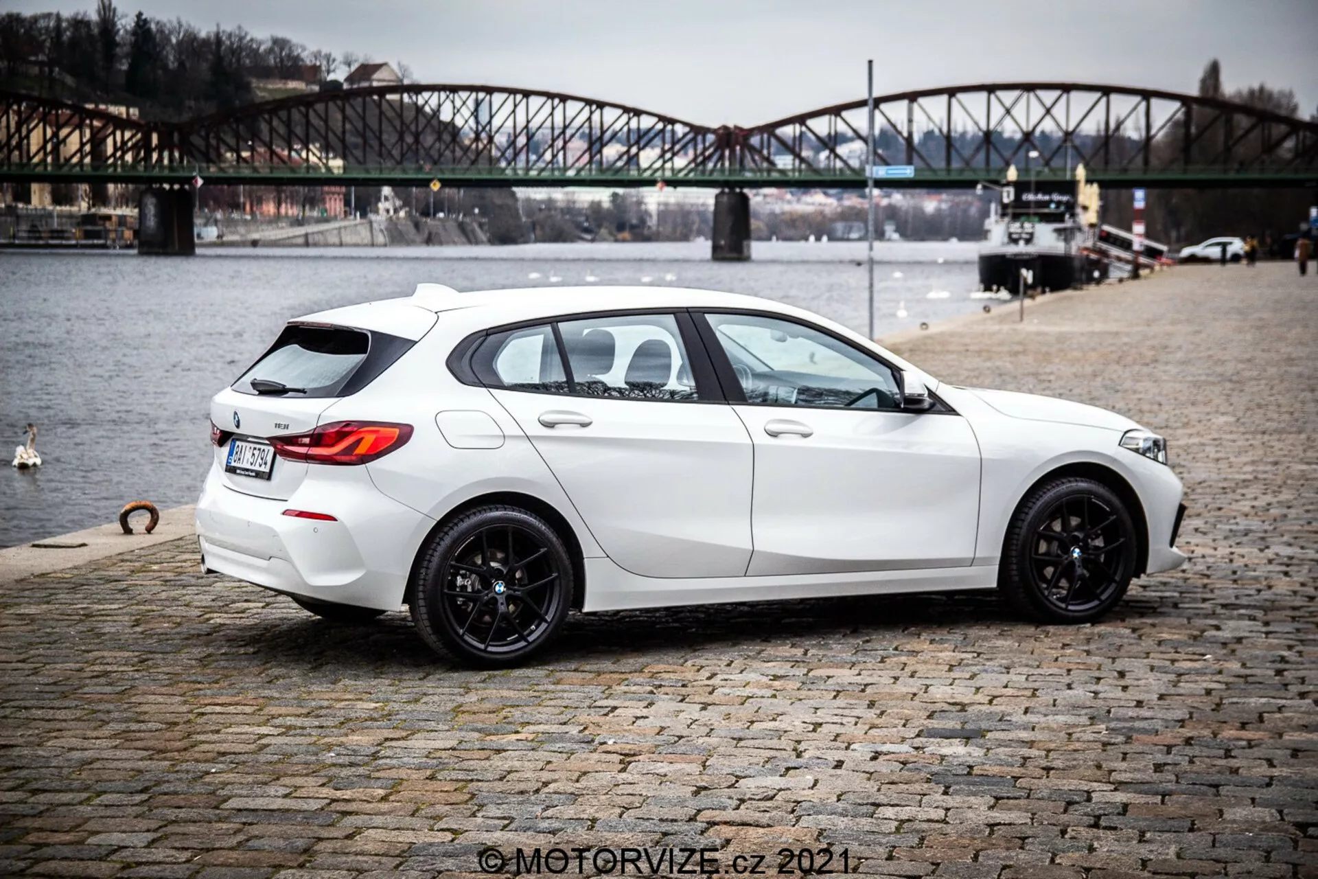 Imagem de um hatchback BMW 1er-Reihe 116i branco de 2019 visto de uma perspectiva traseira de três quartos, destacando seu design moderno e elegante com postura esportiva, rodas de liga leve pretas de múltiplos raios, janelas levemente escurecidas e características práticas como descongelador e limpador do vidro traseiro. O carro exibe linhas e contornos nítidos, sistema de escape duplo, discreto spoiler traseiro no teto, câmera de ré e sensores de estacionamento. Está estacionado em uma superfície de paralelepípedos perto de um corpo d'água com uma ponte urbana ao fundo, sugerindo um contexto de estilo de vida ao ar livre.