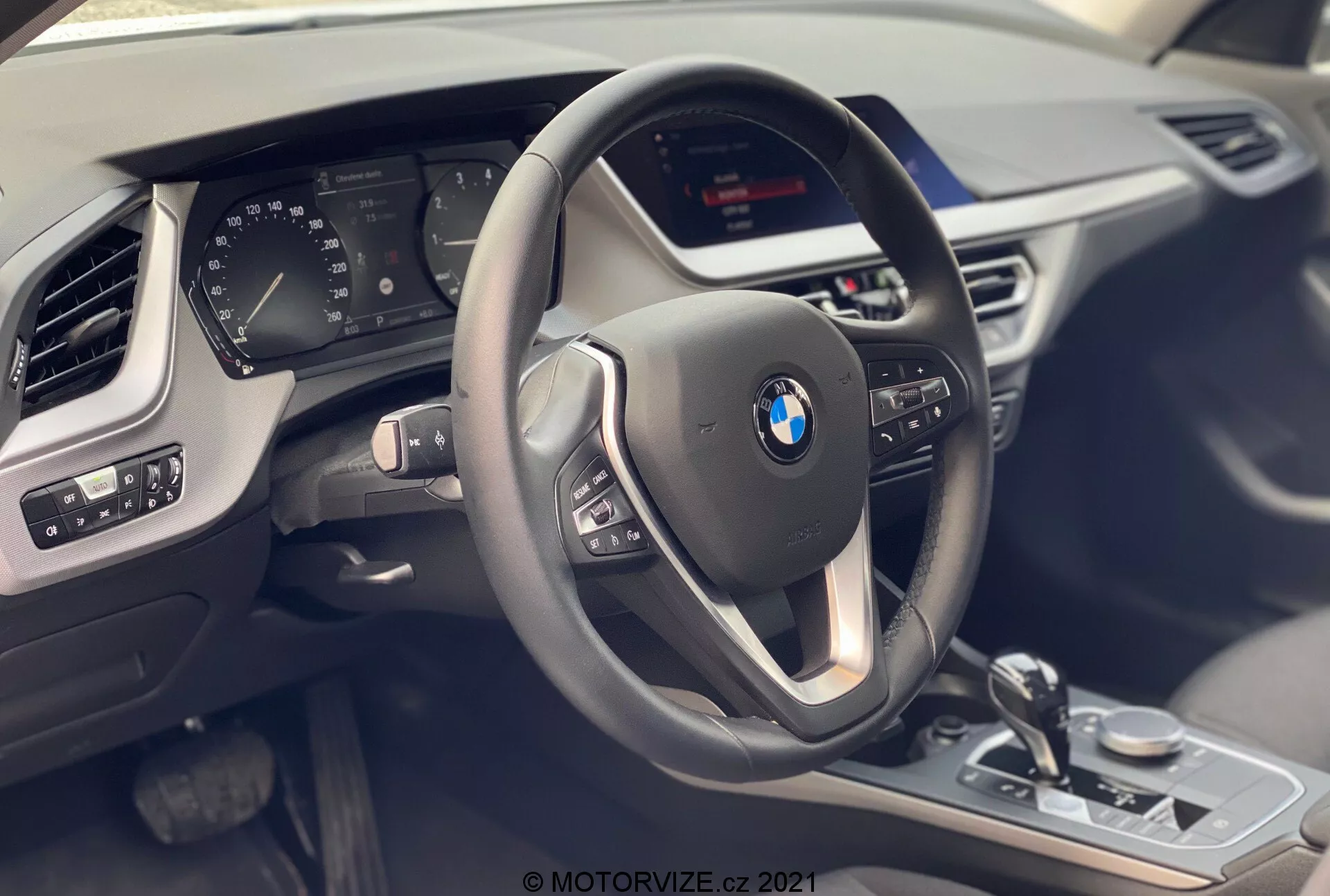 Vista interior do BMW Série 1 2019 (BMW 116i) a partir do banco do passageiro, destacando o cockpit do motorista com um volante moderno de três raios com botões multifuncionais e revestimento de couro com detalhes de costura contrastante. Atrás do volante há um painel de instrumentos digital parcialmente visível que mostra várias métricas do veículo. A consola central abriga uma tela de infotainment não visível e controles visíveis como a alavanca de seletor de marchas, o botão de controle iDrive, o seletor de modos de condução e os controles dos sensores de estacionamento, todos com acabamentos metálicos e acabamento preto brilhante. O painel da porta do motorista mostra ajustes de espelhos e controles de janelas elétricas. Também é visível parte da ventilação lateral do lado do passageiro e a textura do painel, enfatizando luxo, tecnologia e design moderno.