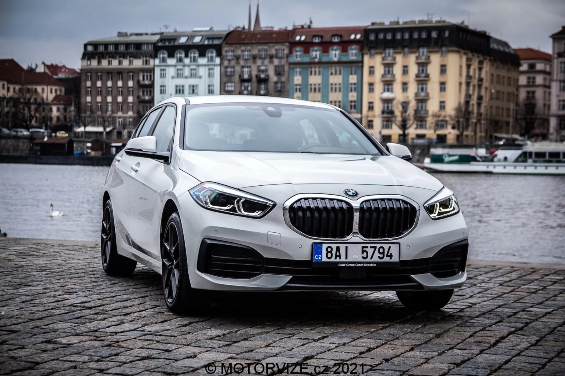 Imagem mostrando um BMW 1er-Reihe 2019 (BMW 116i) de uma perspectiva frontal de três quartos, destacando os contornos e elementos de design do sedã de luxo moderno. O carro está localizado ao ar livre em uma encosta de paralelepípedos perto de um corpo d'água, sob um céu nublado. As características proeminentes incluem uma grade com lamelas verticais, elegantes faróis de LED, luzes de neblina integradas, rodas de liga e perfil aerodinâmico.