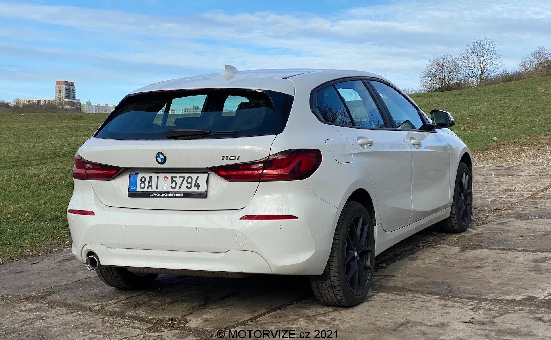 Uma visão de três quartos traseiros de um hatchback BMW 1 Series 118i branco, estacionado em terreno irregular. O carro apresenta luzes traseiras distintivas em forma de 'L', um emblema BMW e um emblema de modelo que diz '118i'. Possui um sistema de escape duplo com um tubo visível no lado direito do para-choques traseiro. O veículo está equipado com rodas de liga preta com múltiplos raios e um limpador de para-brisa traseiro. Um spoiler de teto adiciona ao visual esportivo, e o perfil lateral revela janelas ligeiramente tingidas, dobras na carroçaria e puxadores e espelhos laterais que combinam com o esquema de cores do carro. O ângulo baixo da câmera enfatiza a postura do carro e o design das rodas.