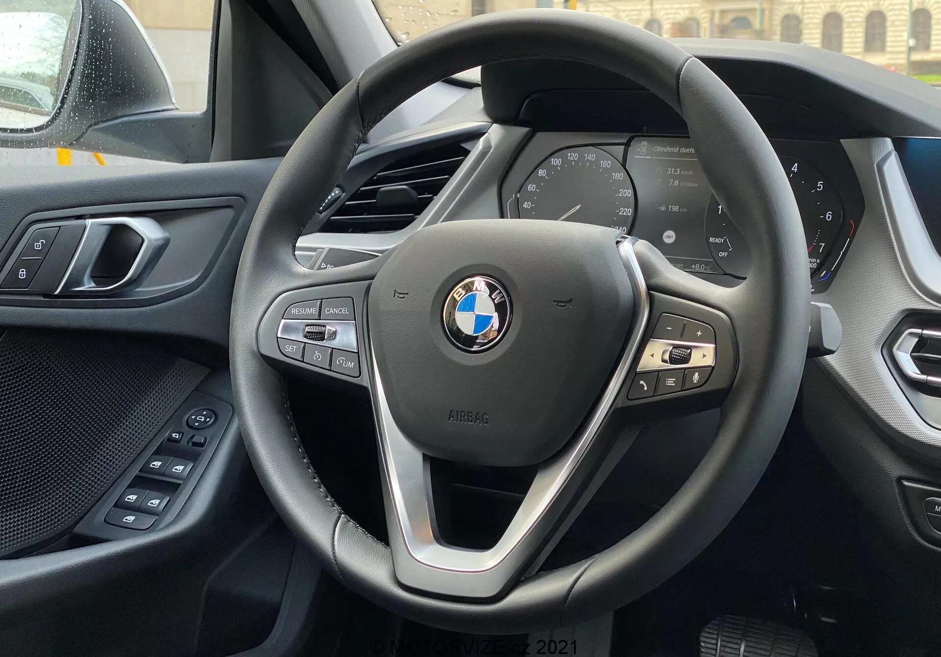 Vista interior de um BMW Série 1 2019 (BMW 116i) a partir do assento do motorista, focando no volante moderno de três raios da BMW com botões de controle para multimídia, telefone e controle de cruzeiro, e o painel de instrumentos digital que exibe métricas essenciais do veículo como velocidade e rpm. A borda superior da consola central é visível ao fundo, e à esquerda vê-se parte da porta do motorista com controlos elétricos de conforto. O painel de instrumentos e o painel da porta exibem uma combinação de materiais suaves ao toque e de alta qualidade. O emblema BMW e o distintivo iPERFORMANCE são proeminentemente exibidos no volante, sugerindo eficiência ou tecnologia híbrida.