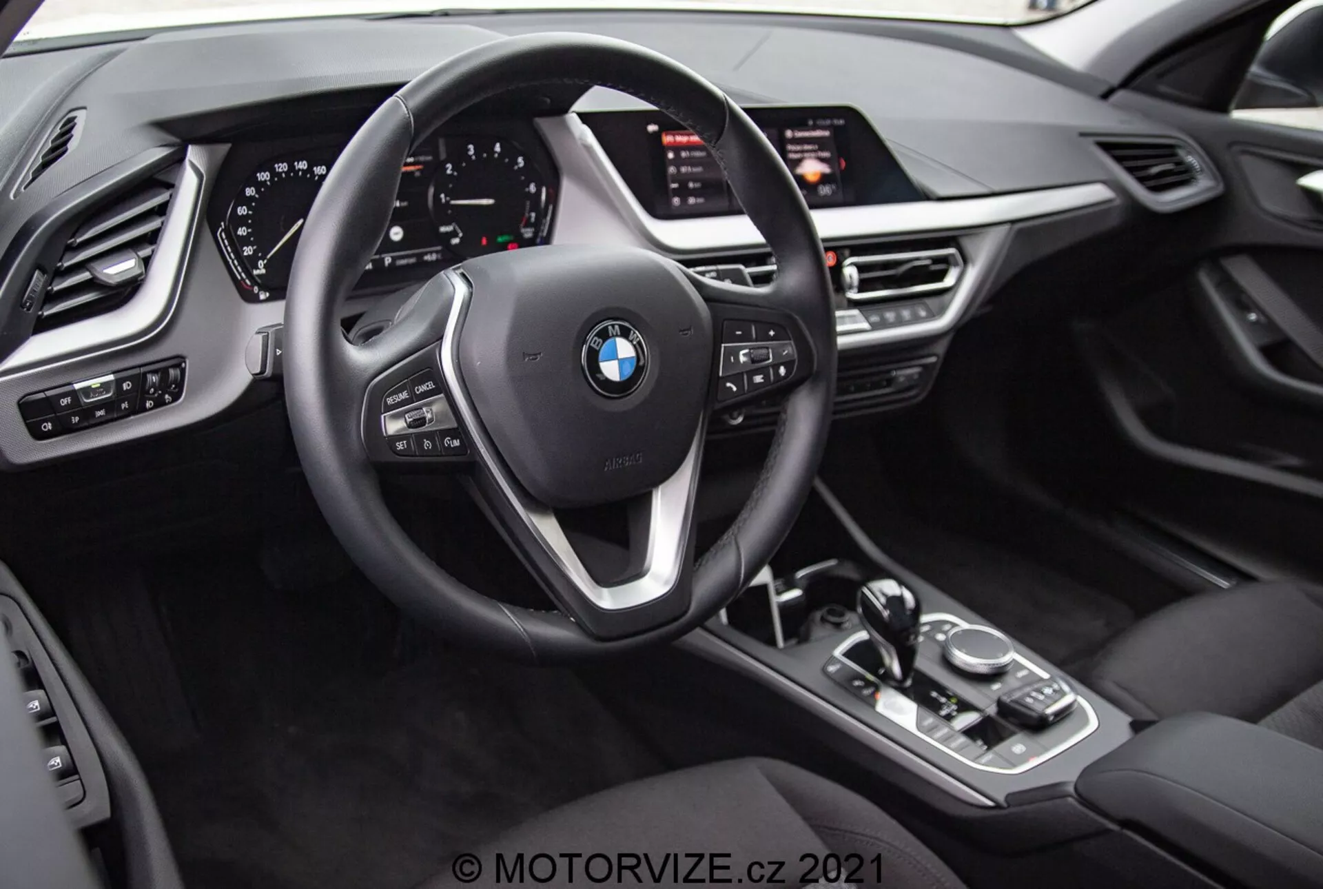 Vista interna do BMW 1er-reihe 2019 (BMW 116i) a partir da perspectiva do motorista. A imagem destaca o emblema da BMW no volante de três raios com botões multifuncionais e paddle shifters atrás do volante. Atrás do volante há um painel de instrumentos totalmente digital com mostradores iluminados e vários displays informativos. A consola central possui uma alavanca de mudança de marchas automática, botão de controlo iDrive, botões para modos de condução e um grande ecrã de infoentretenimento inclinado para o condutor. O design interior inclui texturas semelhantes a couro, acentos metálicos e controles adicionais para faróis, janelas e ajustes dos retrovisores com funções de memória para o assento do motorista.