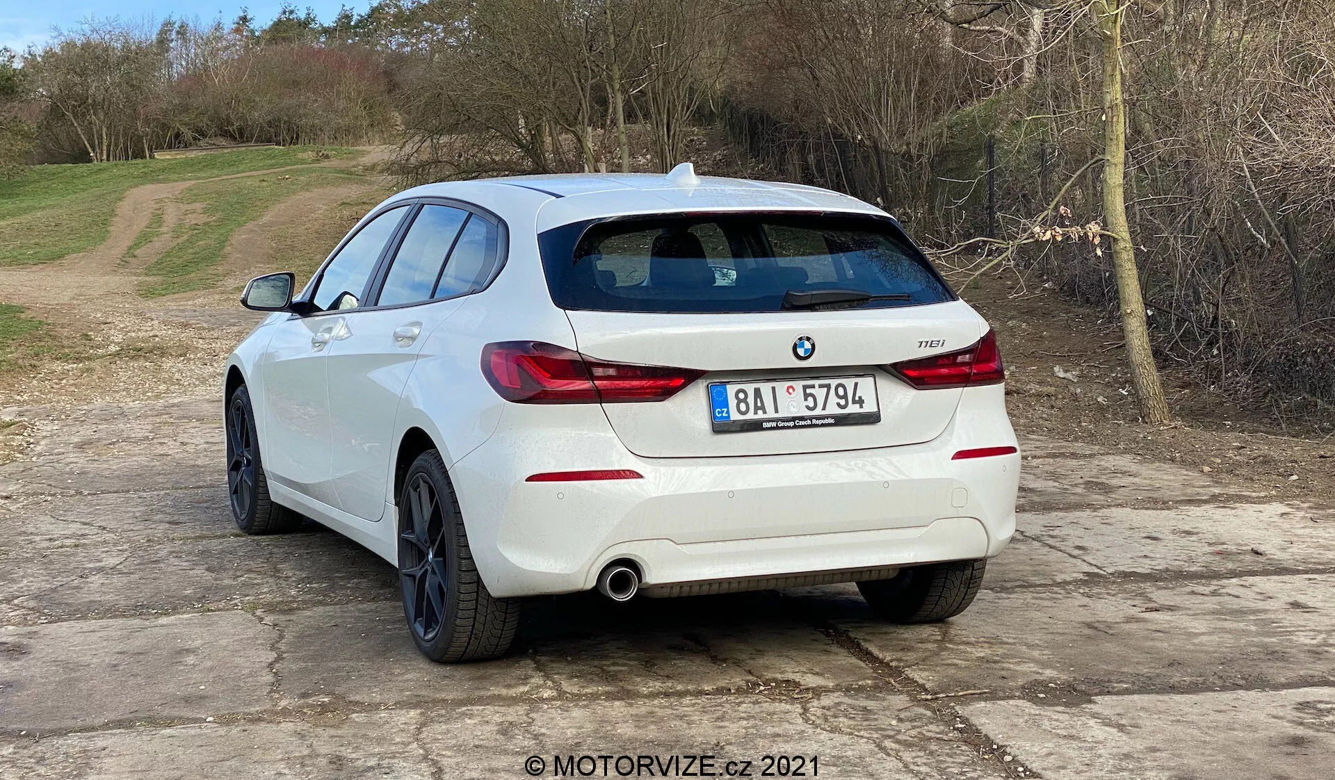 A imagem mostra um BMW 1er-reihe 2019 (BMW 116i) branco hatchback visto de um ângulo traseiro três quartos. A parte traseira do veículo, incluindo as luzes traseiras horizontalmente orientadas, o sistema de escape duplo e os sensores de estacionamento integrados no para-choque traseiro, são claramente visíveis. O emblema da BMW e o emblema do modelo estão na tampa traseira. O carro está situado em uma superfície de cascalho com um fundo natural e rural. A imagem sugere temas automotivos, como design de veículos, modelos contemporâneos, conteúdo específico da BMW, sistemas de escape e tecnologia de assistência ao estacionamento. A presença da marca d'água MOTORVIZE.cz 2021 indica uma origem em uma revisão ou publicação automotiva.