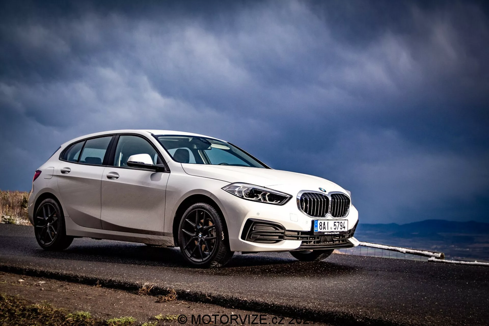 BMW Série 1 2019 (BMW-116i) capturado em uma vista frontal de três quartos, exibindo seu perfil frontal e lateral. O carro, terminado em branco brilhante com acessórios pretos contrastantes, como capas de espelho, moldura de janela e rodas de múltiplos raios, está posicionado no lado esquerdo da moldura contra um fundo de uma estrada asfaltada que se estende à distância e céu nublado. As características visíveis incluem faróis detalhados com elementos circulares, grade de rim proeminente, parachoque dianteiro com entradas de ar e potenciais sensores de estacionamento.