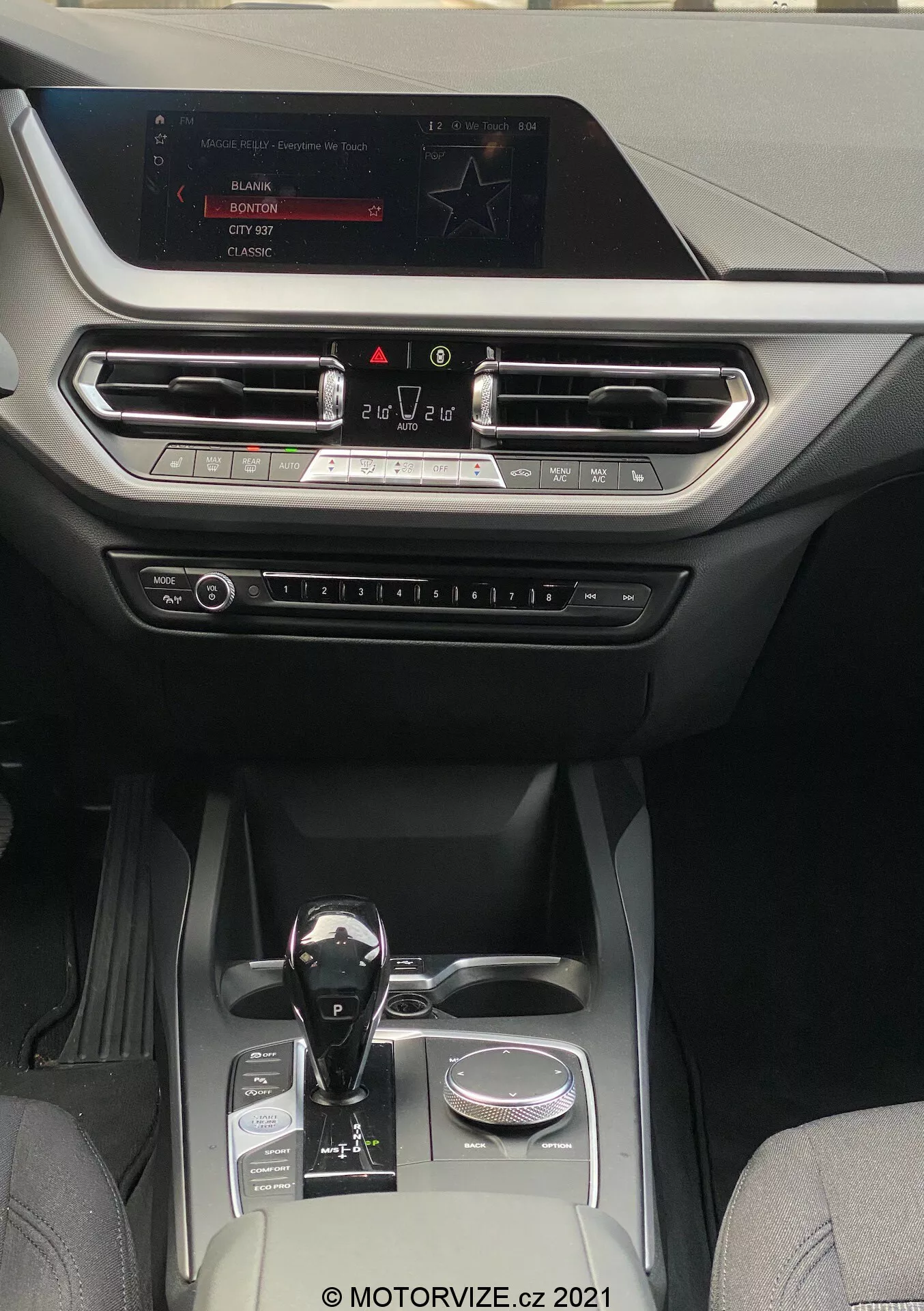 Vista do interior de um BMW 1er-reihe 2019 (BMW-116i), mostrando um habitáculo moderno e luxuoso com foco na consola central e área do painel de instrumentos. A perspectiva é da área dos assentos traseiros, olhando para a frente em direção à tela de entretenimento montada no topo do painel. A tela mostra opções de rádio, mídia e configurações do carro. Abaixo da tela, há saídas de ventilação alinhadas horizontalmente com detalhes metálicos e um acabamento decorativo que se estende por toda a largura do painel. Abaixo das saídas de ventilação, são visíveis botões físicos e comandos para controle climático e outras configurações. A alavanca de câmbio automático tem um acabamento preto brilhante com detalhes metálicos, posicionada ao lado de um botão rotativo. A consola central inclui dois porta-copos, um pequeno compartimento de armazenamento e um elemento decorativo prateado, indicando um habitáculo bem equipado que combina couro e tecidos sintéticos de alta qualidade, adequado para categorias de veículos de médio a alto padrão focados em tecnologia e conforto.