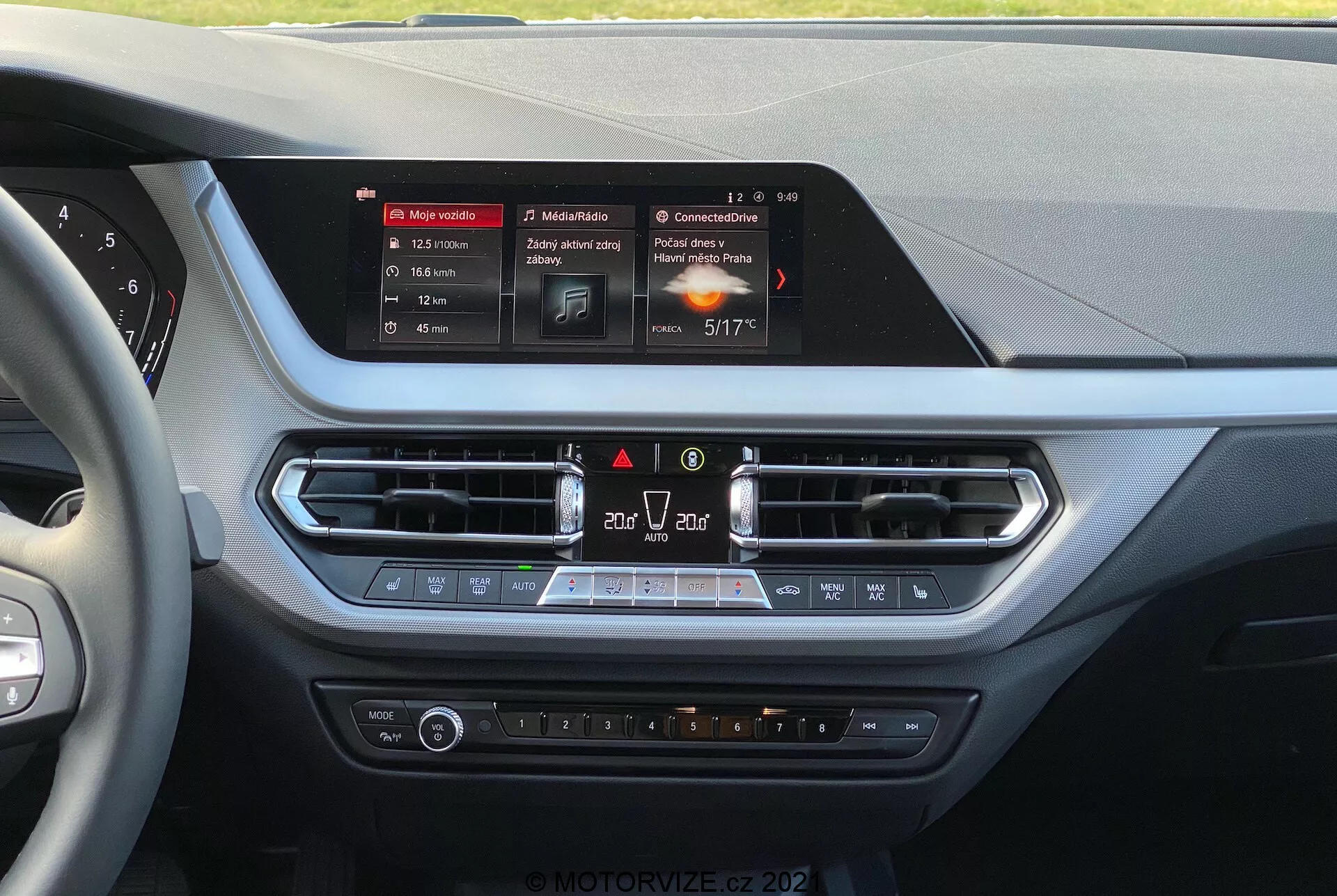 Vista interior de um BMW série 1 de 2019 (BMW-116i) a partir da perspectiva do condutor, mostrando o console central do painel. No centro está o ecrã táctil retangular e iluminado do sistema de infotainment, exibindo navegação, multimídia e detalhes climáticos. Abaixo da tela, há aberturas de ar com acabamento metálico, controles de emergência e recirculação de ar, seguidos por um painel de controle climático com botões de temperatura, velocidade do ventilador e assentos aquecidos. Os materiais apresentam superfícies macias ao toque e detalhes cromados, sugerindo foco no conforto e conveniência do condutor e do passageiro da frente.