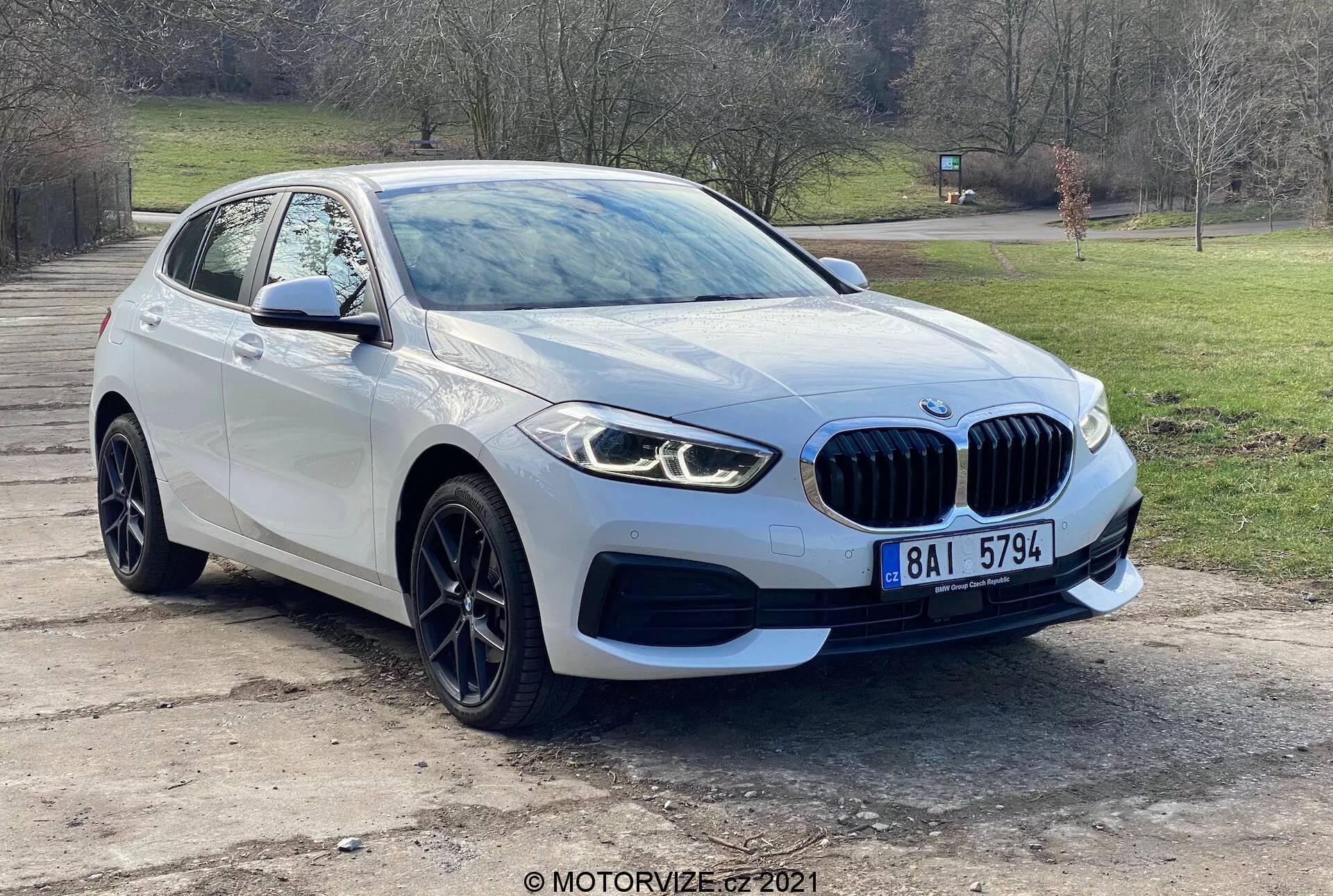 Veículo moderno BMW 116i da Série 1 de 2019 fotografado de uma perspectiva frontal de três quartos, mostrando a grelha dianteira com emblema, elegantes faróis LED, luzes de nevoeiro, para-choques dianteiro e linhas aerodinâmicas do capô que conduzem ao para-brisas. A roda e o pára-choques do lado do condutor, juntamente com o espelho lateral com indicadores de direção integrados, destacam o design avançado e a tecnologia de sensores de estacionamento do veículo. Capturado ao ar livre com fundo de árvores e grama, o carro possui rodas de liga leve e uma grelha distinta em forma de rim, indicando luxo e desempenho.