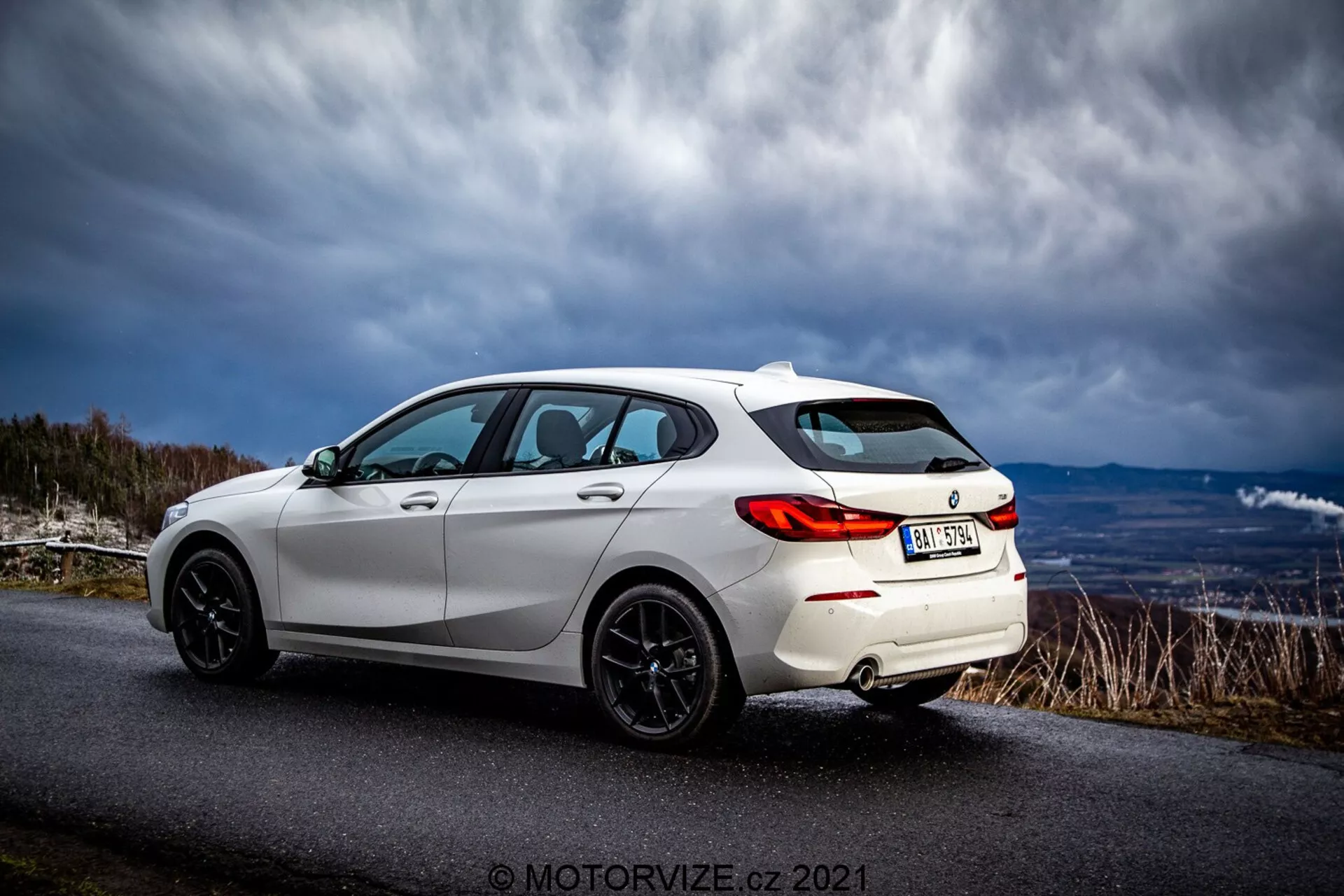Um hatchback moderno branco BMW 116i 2019 capturado em uma vista traseira de três quartos. As características esportivas do exterior incluem luzes traseiras LED, rodas de liga leve pretas de múltiplos raios, um spoiler traseiro, janelas com insulfilme e linhas elegantes da carroceria que destacam seu design aerodinâmico. O veículo está estacionado em uma estrada asfaltada em uma área rural ou montanhosa sob um céu dramático.