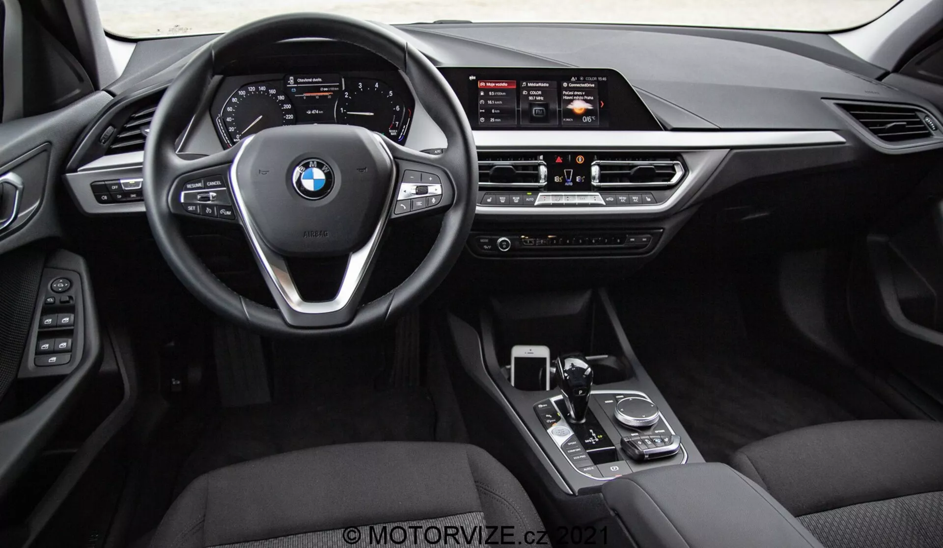 Vista interna de um BMW Série 1 2019 (BMW 116i) tirada dos bancos traseiros, mostrando o painel frontal completo, volante com logótipo BMW e botões de controle, e console central. Recursos visíveis incluem o amplo ecrã de infoentretenimento, controles manuais de climatização, saídas de ar, alavanca de câmbio, tecla rotativa e touchpad. O interior possui acabamentos em couro preto ou couro sintético com elementos metálicos e brilhantes, enfatizando o luxo e a tecnologia avançada da interface do condutor.