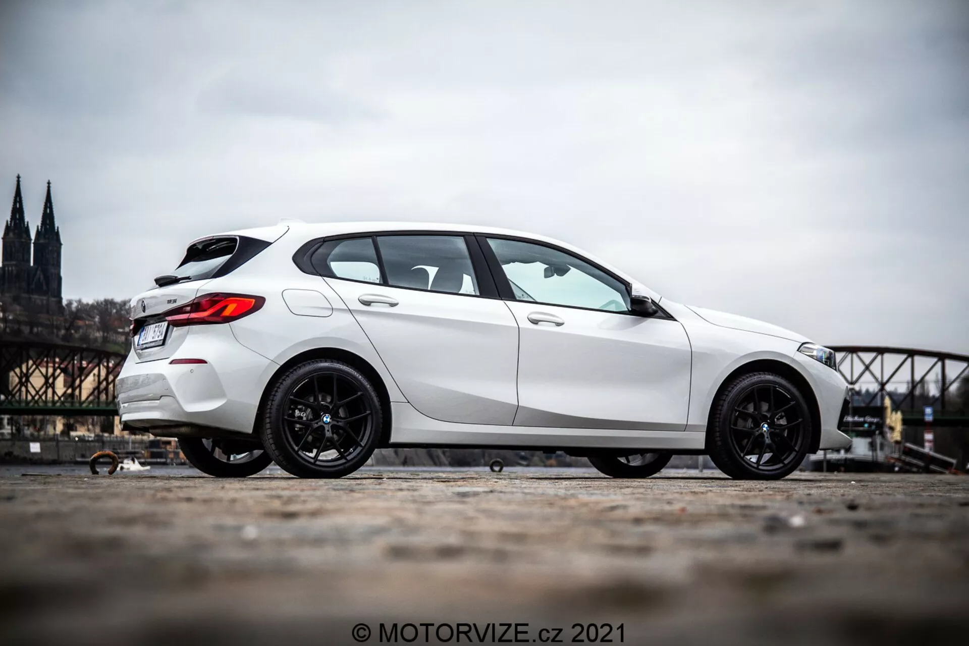 Um hatchback BMW Série 1 (BMW 116i) branco de 2019 fotografado de um ângulo lateral de perfil, mostrando o lado do veículo de pouco antes da roda dianteira até a traseira. O carro está posicionado no lado esquerdo da imagem com um fundo urbano histórico desfocado que inclui uma catedral e uma ponte. A imagem destaca as rodas esportivas de cor escura do carro e a postura dominante do veículo devido à perspectiva de nível de solo inferior e à inclinação da câmera para cima. A foto é tirada ao ar livre durante o dia, revelando detalhes da estética do carro à luz natural do dia.