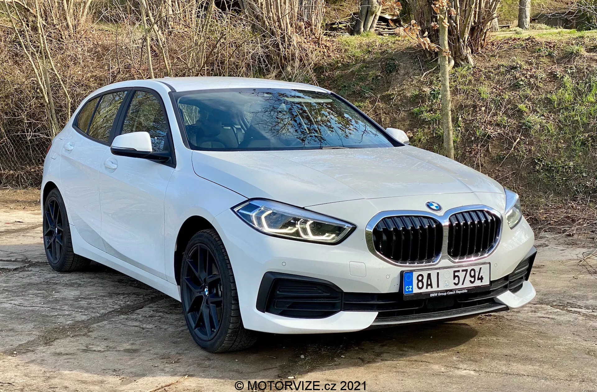 BMW 116i Série 1 2019 em branco, capturado de um ângulo de três quartos frontais, destacando sua proeminente grade frontal em forma de rim, faróis LED angulares, rodas pretas elegantes com pneus de perfil baixo e linhas aerodinâmicas da carroceria. Posicionado ao ar livre em uma superfície plana com vegetação natural ao fundo, a foto destaca o design moderno e os recursos esportivos do carro, incluindo sensores de estacionamento e espelhos laterais com indicadores de direção integrados.