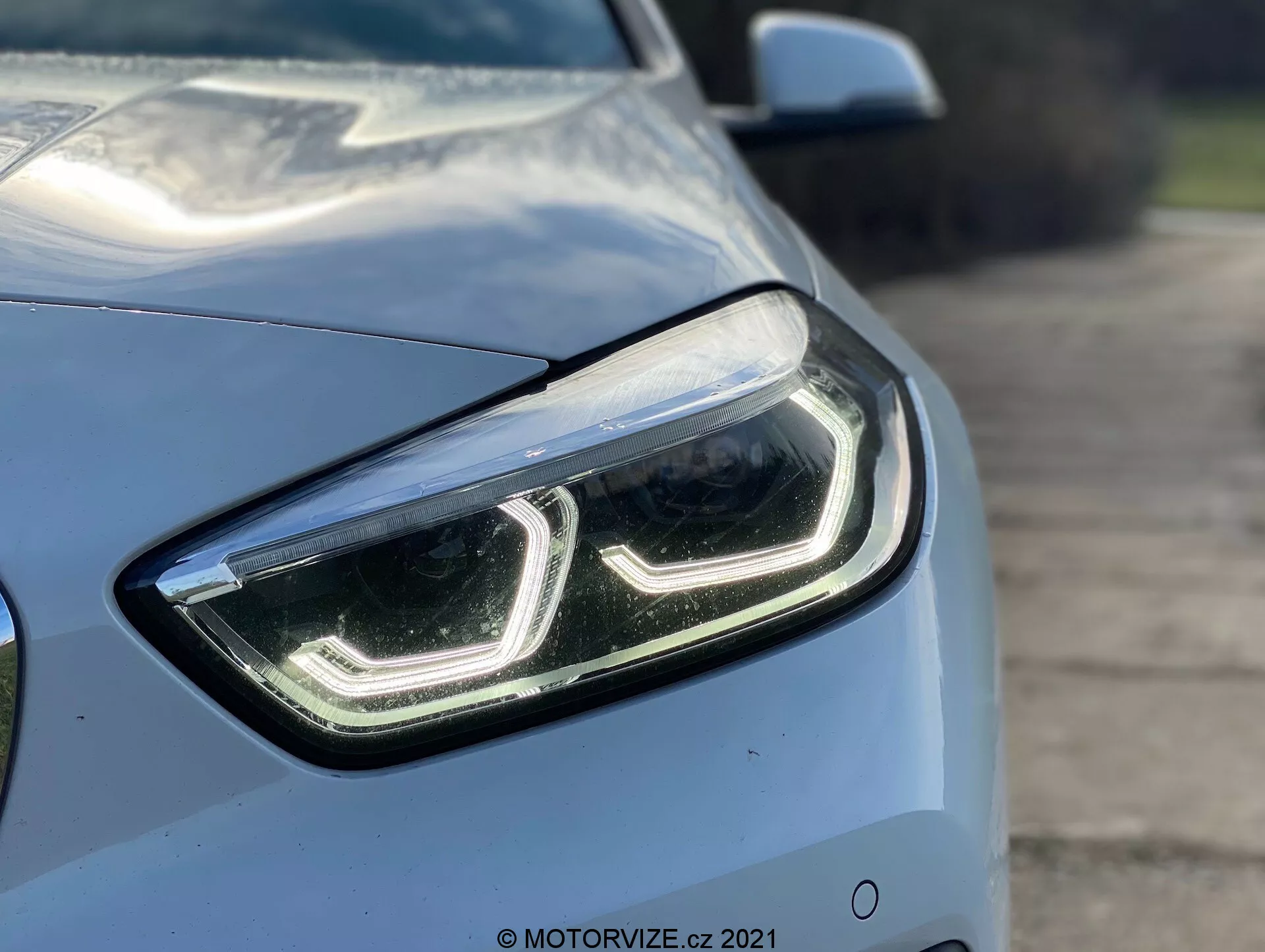 Close-up da BMW Série 1 2019 (BMW 116i) a partir de um ângulo de frente, destacando o lado esquerdo e focando no design moderno e elegante do farol dianteiro esquerdo com luzes diurnas em LED e cobertura de lente transparente. As linhas aerodinâmicas do capô e do para-lama são parcialmente visíveis, mostrando a cor metálica clara e o acabamento brilhante do carro. Um fundo desfocado enfatiza as características tecnológicas avançadas do farol, sugerindo lâmpadas LED ou HID, possivelmente equipadas com tecnologia de iluminação adaptativa.