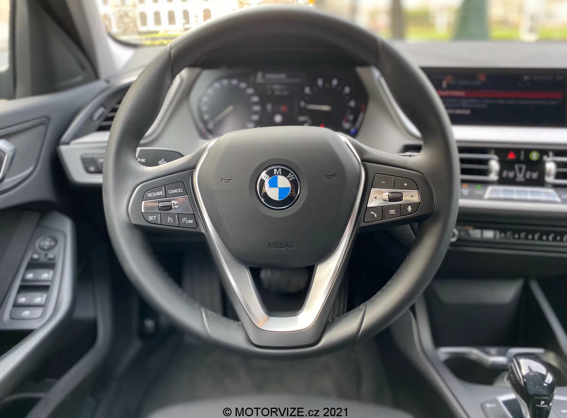 Perspectiva do assento do motorista de um BMW Série 1 2019 (BMW-116i), focando no volante contemporâneo de três raios com um detalhe metálico no raio inferior e o emblema BMW proeminente no centro. No lado esquerdo do volante, há controles de mídia e comunicação, enquanto o lado direito inclui botões de controle de cruzeiro e uma alavanca de câmbio visível. O fundo mostra um painel de instrumentos parcialmente visível com indicadores de estilo analógico e uma tela de infotainment no console central, sugerindo recursos avançados como navegação GPS e controle de clima.