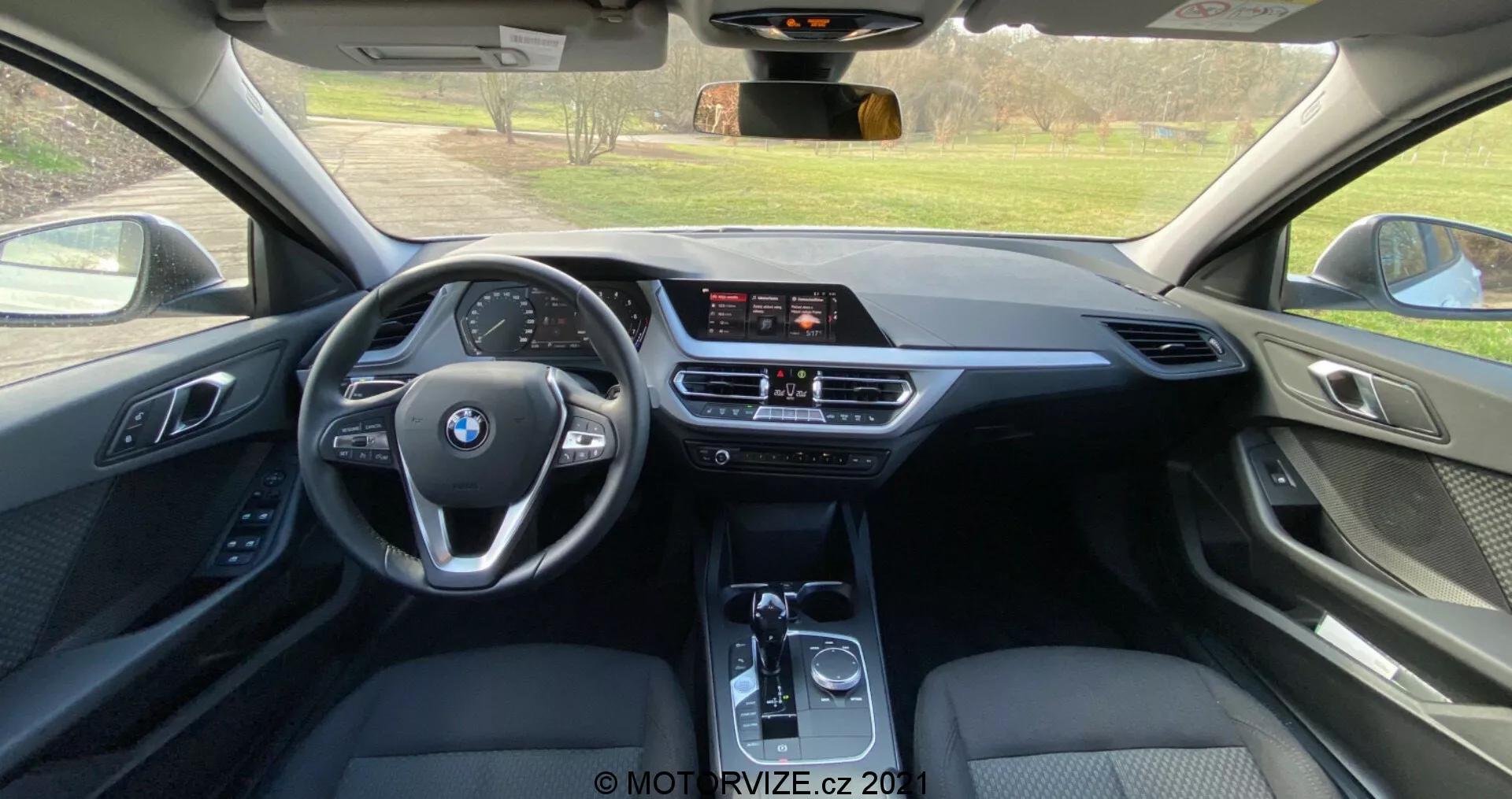 Vista interior do banco traseiro de um BMW Série 1 2019 (BMW-116i), mostrando uma cabine moderna com volante multifuncional de três raios com o logotipo da BMW, tela de infoentretenimento independente, controle de clima de dupla zona e alavanca de câmbio automática. O uso de couro, plásticos macios e detalhes metálicos destaca um nível de acabamento de alta qualidade.