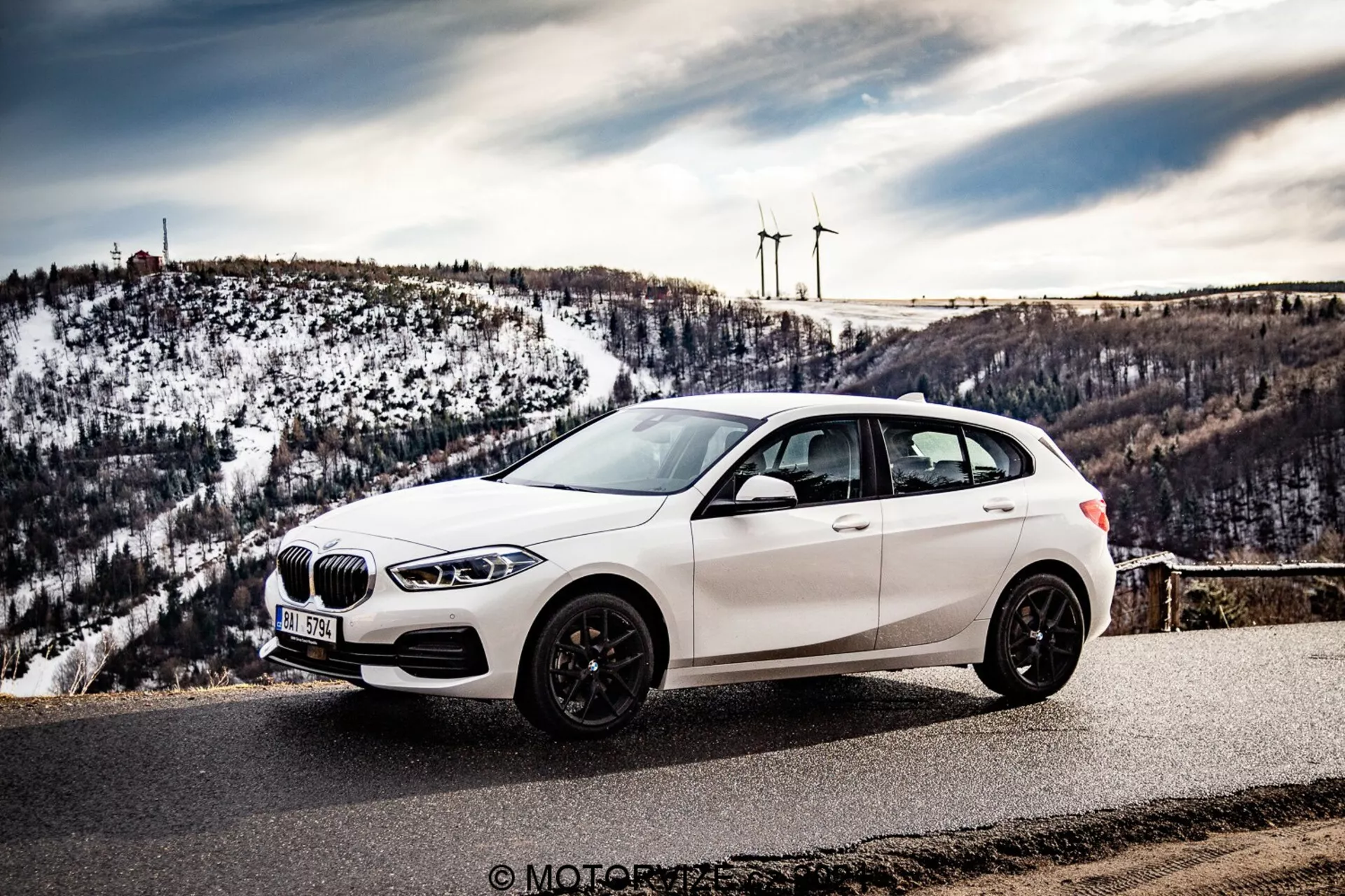 A imagem mostra um BMW 116i hatchback 2019 branco estacionado em um mirante cênico ou estrada de montanha, fotografado de um ângulo frontal lateral que exibe a grade frontal, faróis de LED, roda dianteira, perfil lateral e um vislumbre da seção traseira. A perspectiva é ligeiramente elevada, destacando o teto do carro, que está deslocado para a esquerda, com um fundo de colinas nevadas e turbinas eólicas distantes. O veículo possui uma grade em forma de rim, pneus de perfil baixo em rodas de liga leve de múltiplos raios e rodas de cor escura, espelhos laterais com indicadores de direção integrados e janelas escurecidas. O ambiente sugere clima claro, melhorando a natureza promocional e visualmente atraente da fotografia, potencialmente tirada durante uma viagem de carro ou uma revisão automotiva.