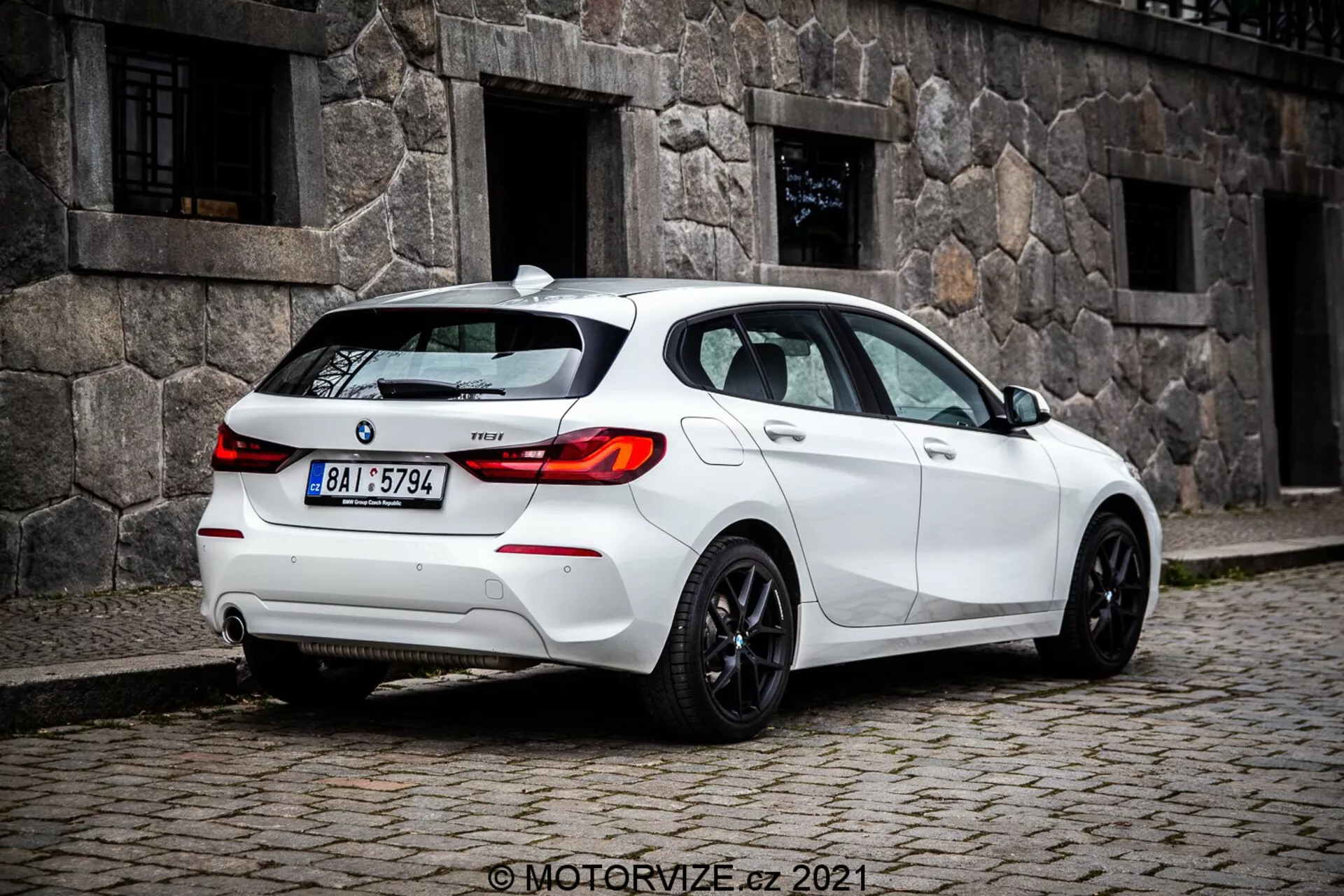 Imagem a média distância no nível do solo de um hatchback branco BMW 1er-série 116i 2019 capturada de um ângulo traseiro de três quartos, mostrando proeminentemente as luzes traseiras com design LED, para-brisa traseiro e rodas esportivas. Os recursos visíveis incluem sensores no para-choque traseiro possivelmente para assistência de estacionamento, uma pequena antena no teto provavelmente para GPS ou rádio por satélite, emblema do modelo no porta-malas, sistema de escape duplo sugerindo desempenho e janelas escurecidas para privacidade ou conforto.